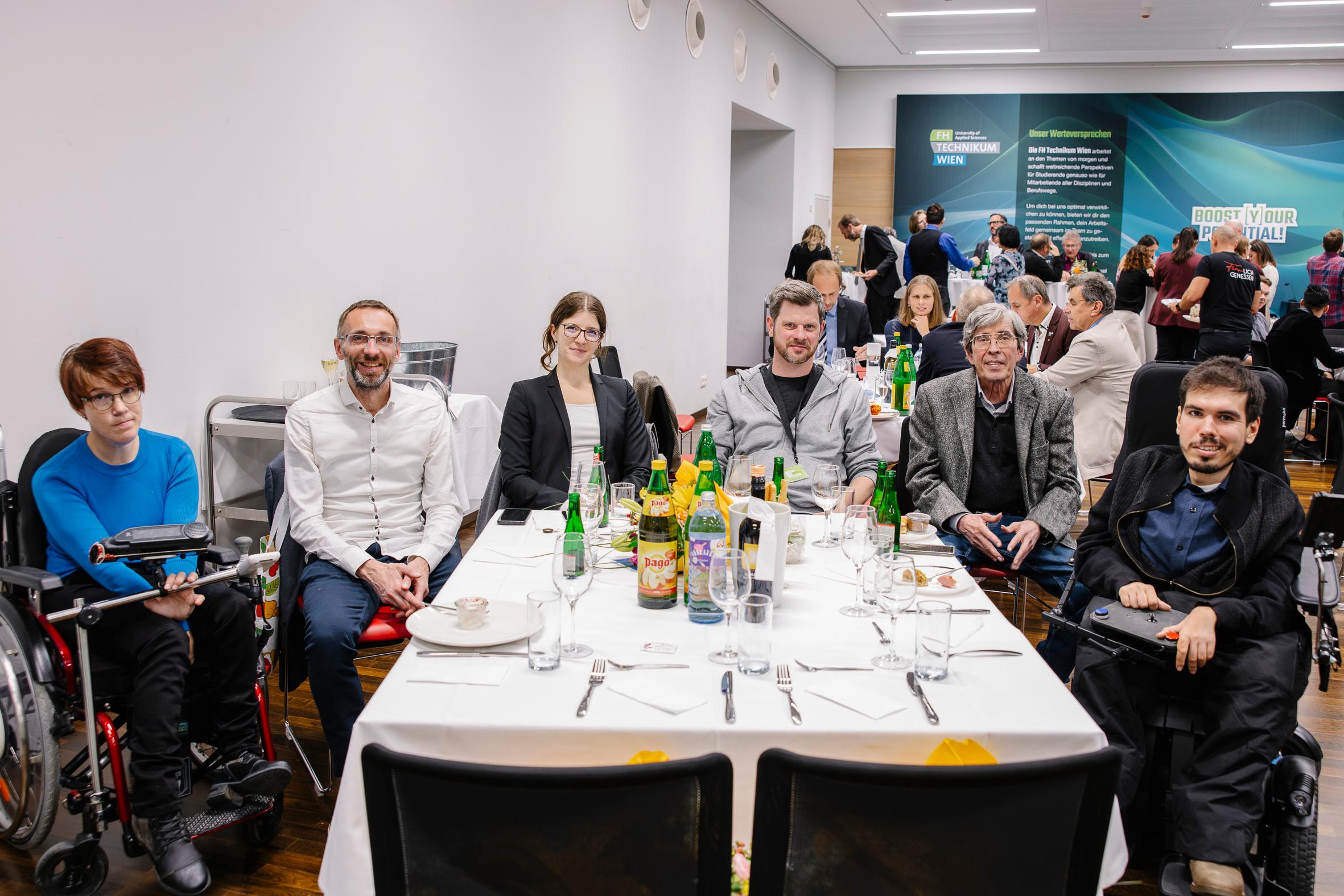 Eine Gruppe von sechs Personen, darunter auch Rollstuhlfahrer, sitzt um einen Tisch, der für eine Mahlzeit bei einer offiziellen Veranstaltung gedeckt ist. Im Hintergrund sind weitere Gäste zu sehen, die sich unterhalten.