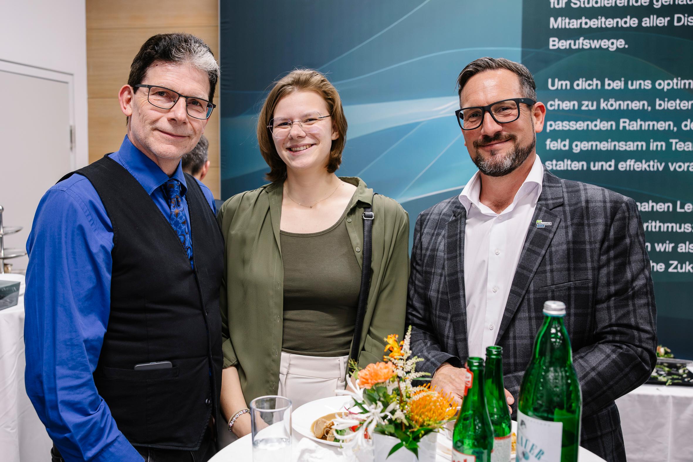 Drei Personen stehen zusammen an einem Tisch mit Getränken und Blumen und lächeln in die Kamera bei einer Indoor-Veranstaltung. Im Hintergrund ist ein blaues und grünes Display mit deutschem Text zu sehen.