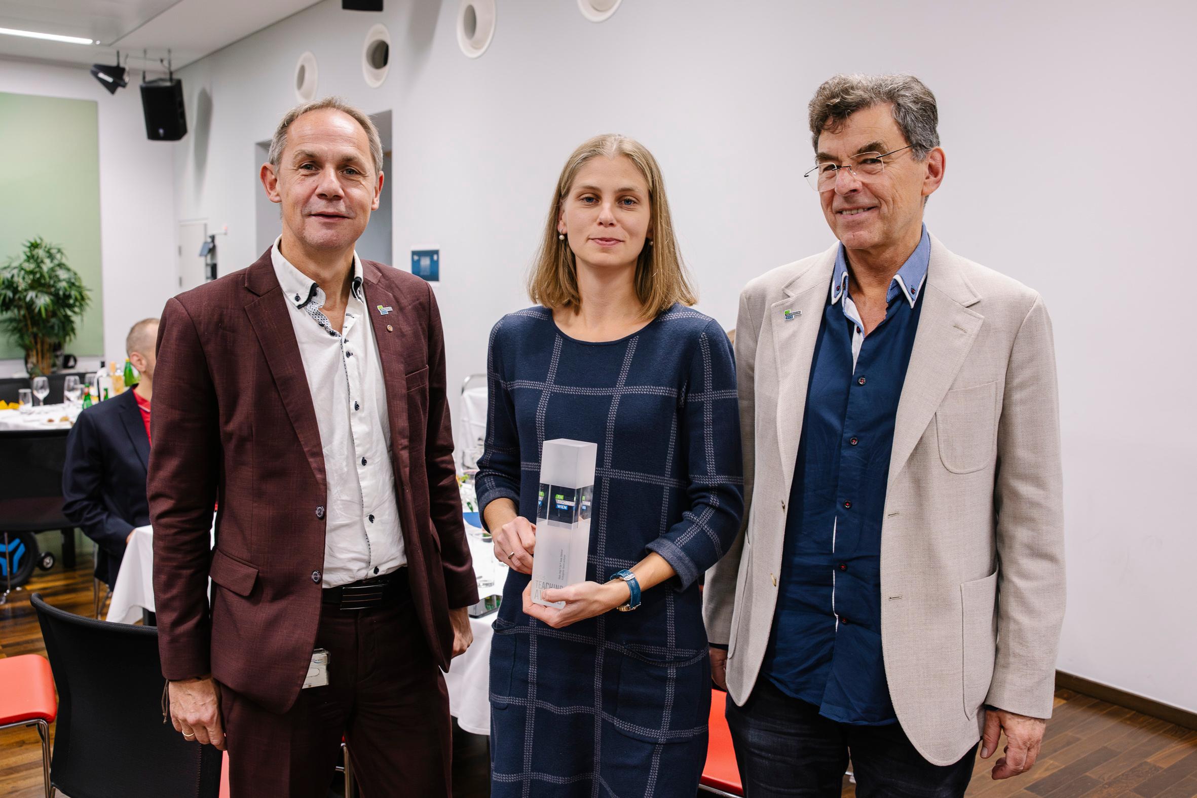 Drei Erwachsene stehen drinnen bei einer Veranstaltung. Die Frau in der Mitte hält eine Trophäe oder Auszeichnung und lächelt in die Kamera. Die beiden Männer an den beiden Seiten schauen ebenfalls in die Kamera. Im Hintergrund sind Tische und Stühle zu sehen.