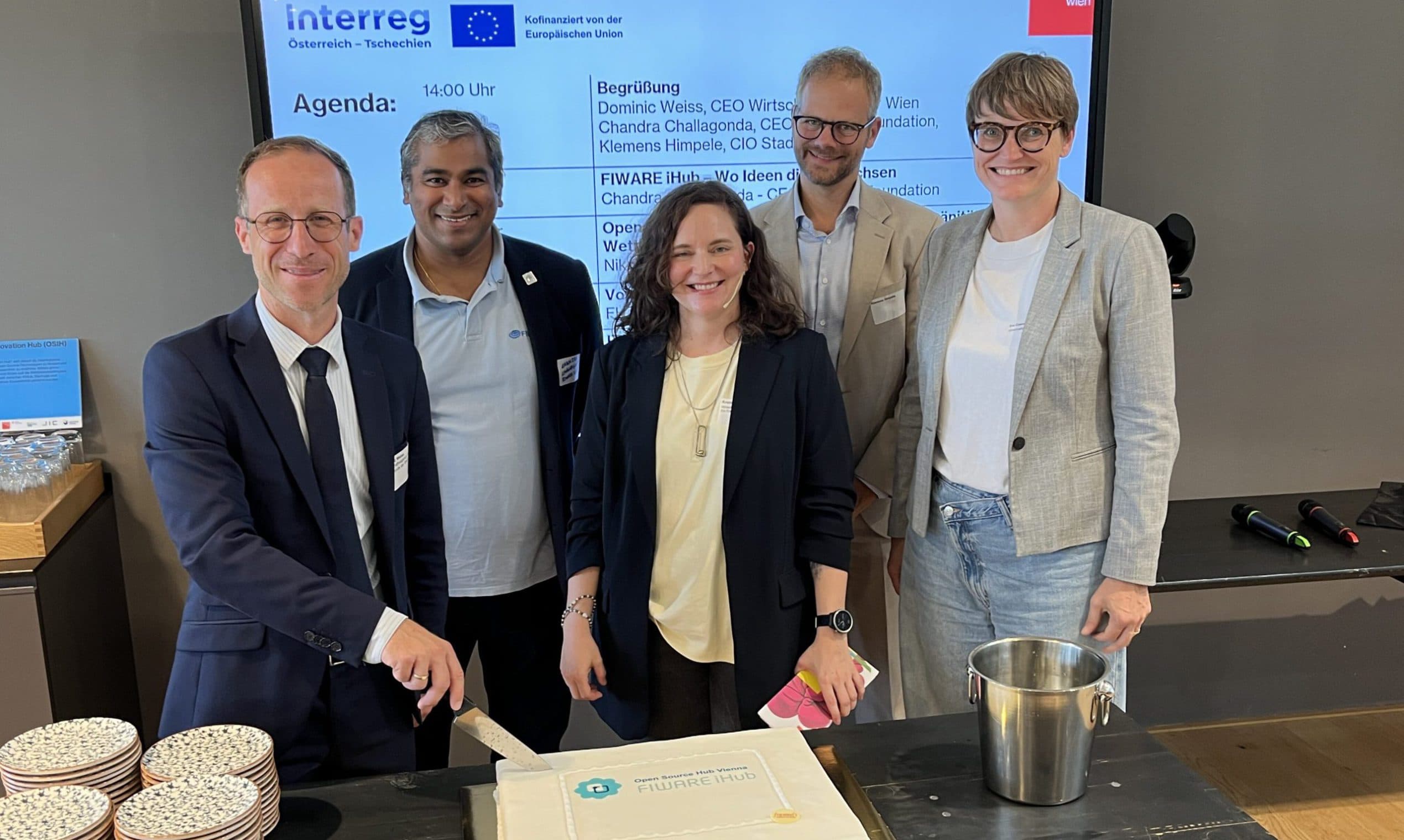 Fünf Personen in Geschäftskleidung lächeln für ein Gruppenfoto, während einer von ihnen einen großen weißen Kuchen auf einem Tisch anschneidet. Links steht ein Stapel von Tellern und Utensilien, und auf einem Bildschirm im Hintergrund wird eine Tagesordnung angezeigt.