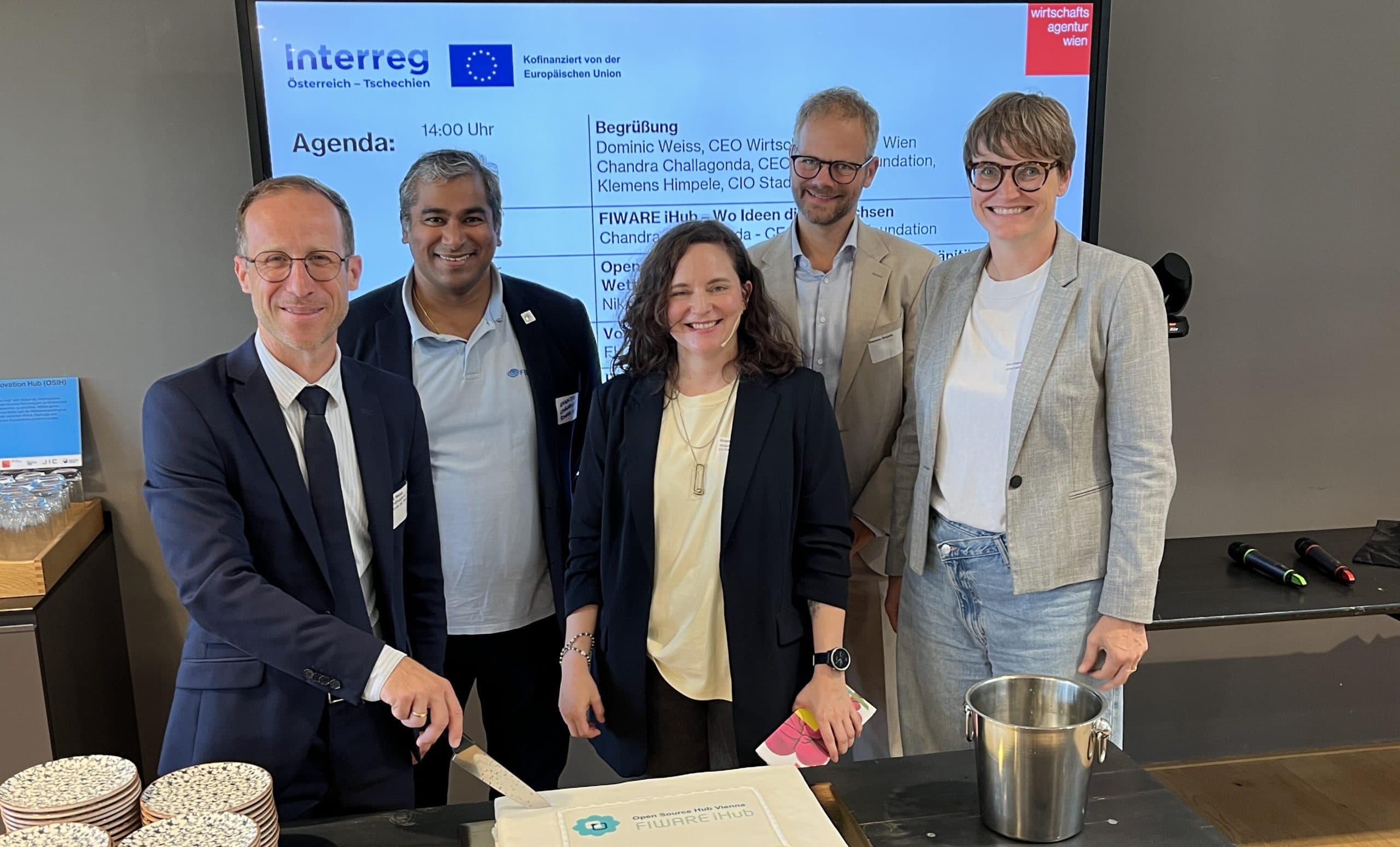 Fünf Personen stehen lächelnd um einen Tisch mit einer großen Torte. Eine Person schneidet die Torte an. Auf dem Tisch stehen die Torte, Teller, Utensilien und ein Metalleimer. Auf einem Bildschirm hinter ihnen werden eine Tagesordnung und Projektinformationen angezeigt.