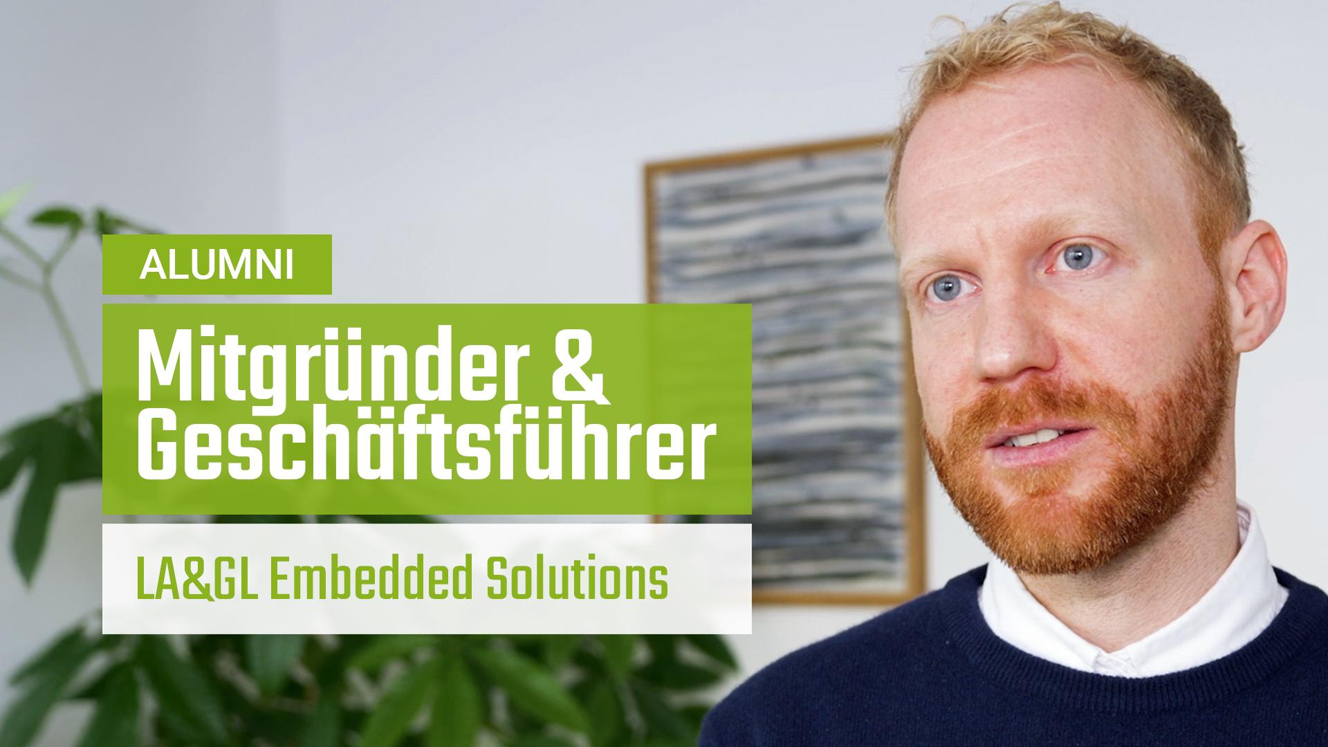 Ein Mann mit roten Haaren und Bart in einem blauen Pullover sitzt in einem Haus. Der überlagerte Text in Deutsch lautet: ALUMNI. Mitgründer & Geschäftsführer. LA&GL Embedded Solutions. Im Hintergrund sind eine Pflanze und ein gerahmtes Bild zu sehen.