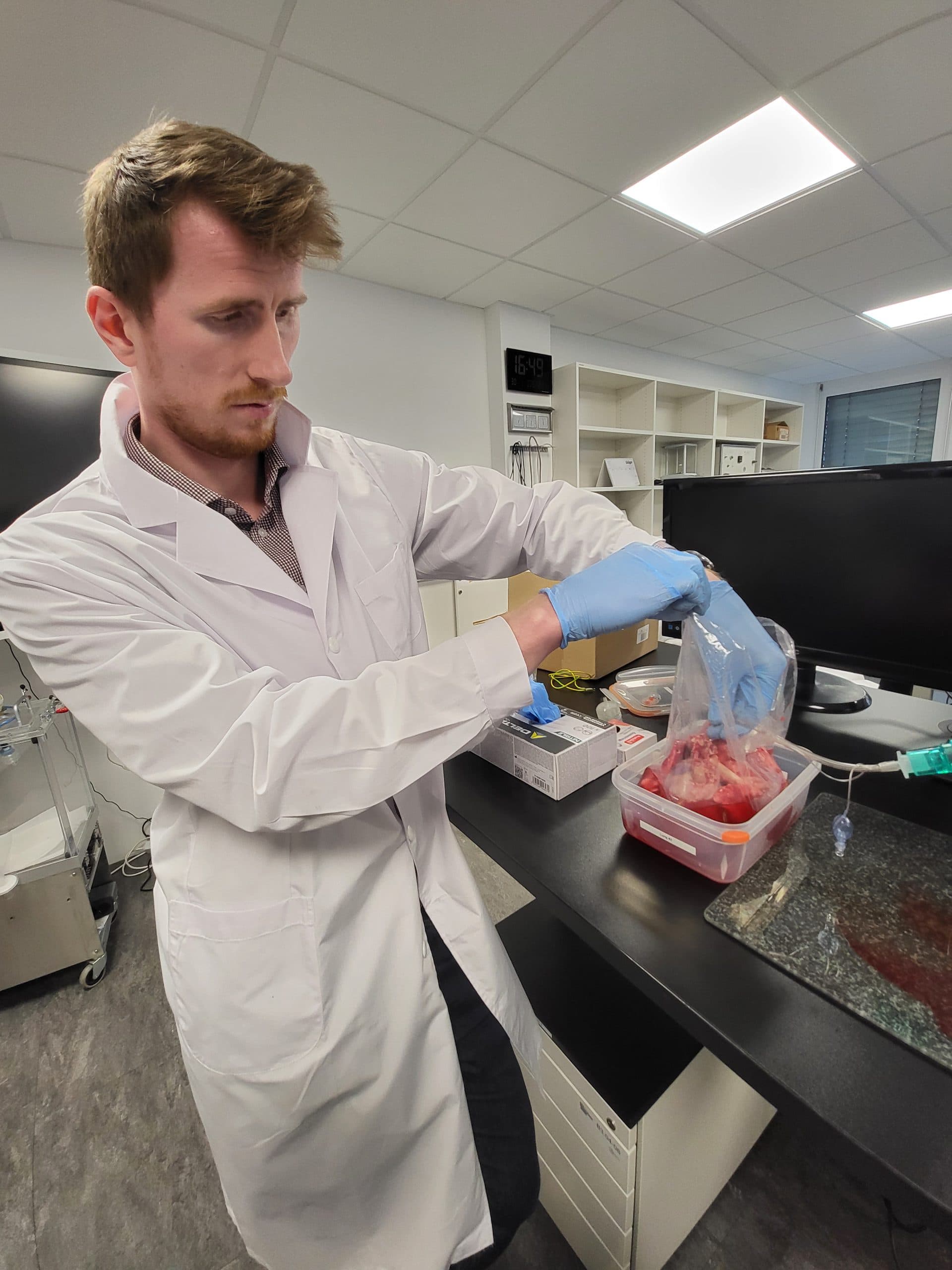 Eine Person in einem Laborkittel und mit blauen Handschuhen hantiert mit einem versiegelten Plastikbeutel, der rotes Material, möglicherweise Gewebe oder Fleisch, enthält, in einer Laborumgebung mit Laborgeräten und Regalen im Hintergrund.