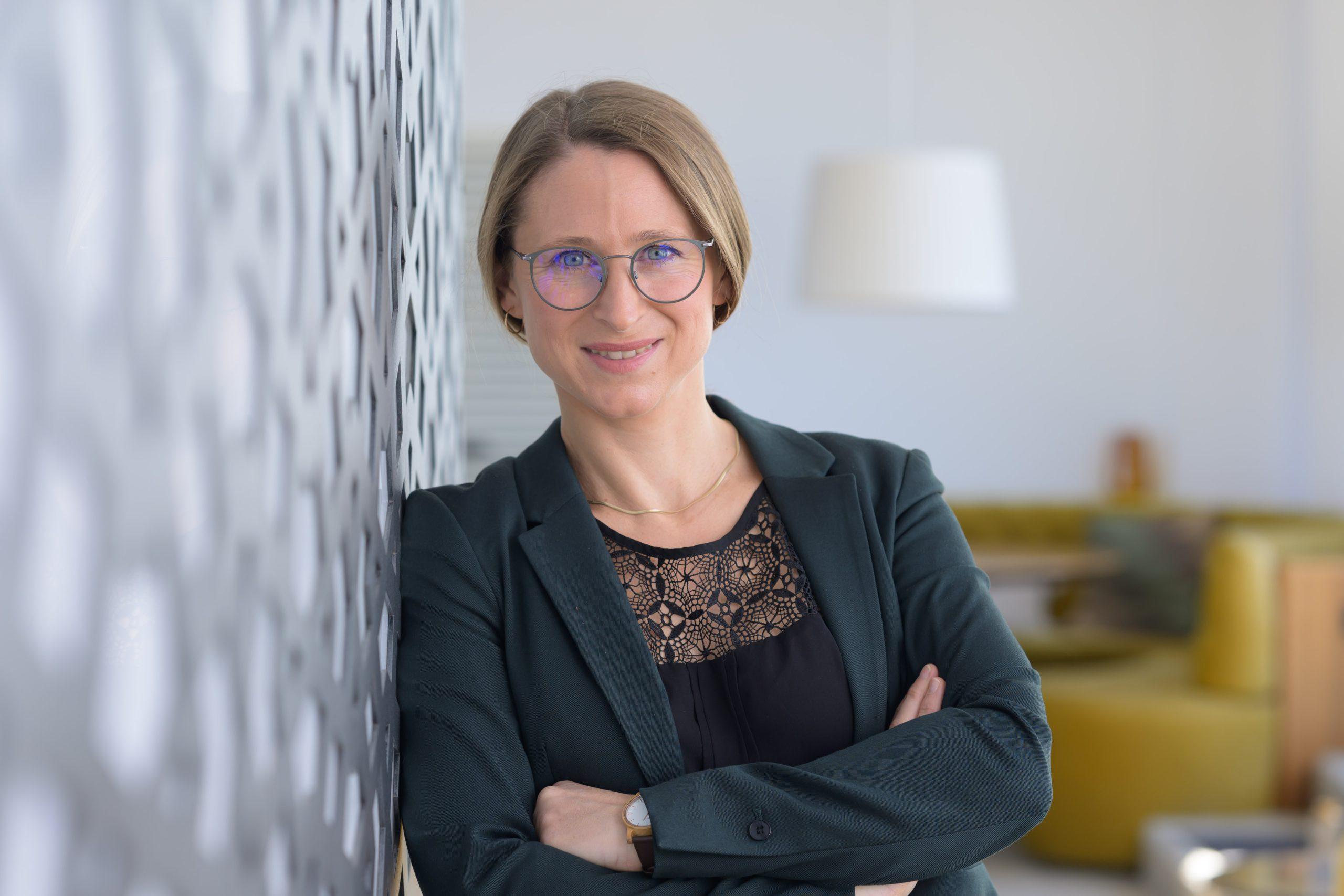 Eine Frau mit hellbraunem Haar und Brille, die einen dunklen Blazer und ein schwarzes Oberteil mit Spitzenverzierung trägt, steht mit verschränkten Armen da und lächelt, während sie sich an eine gemusterte Wand in einem modernen, hell erleuchteten Büroraum lehnt.