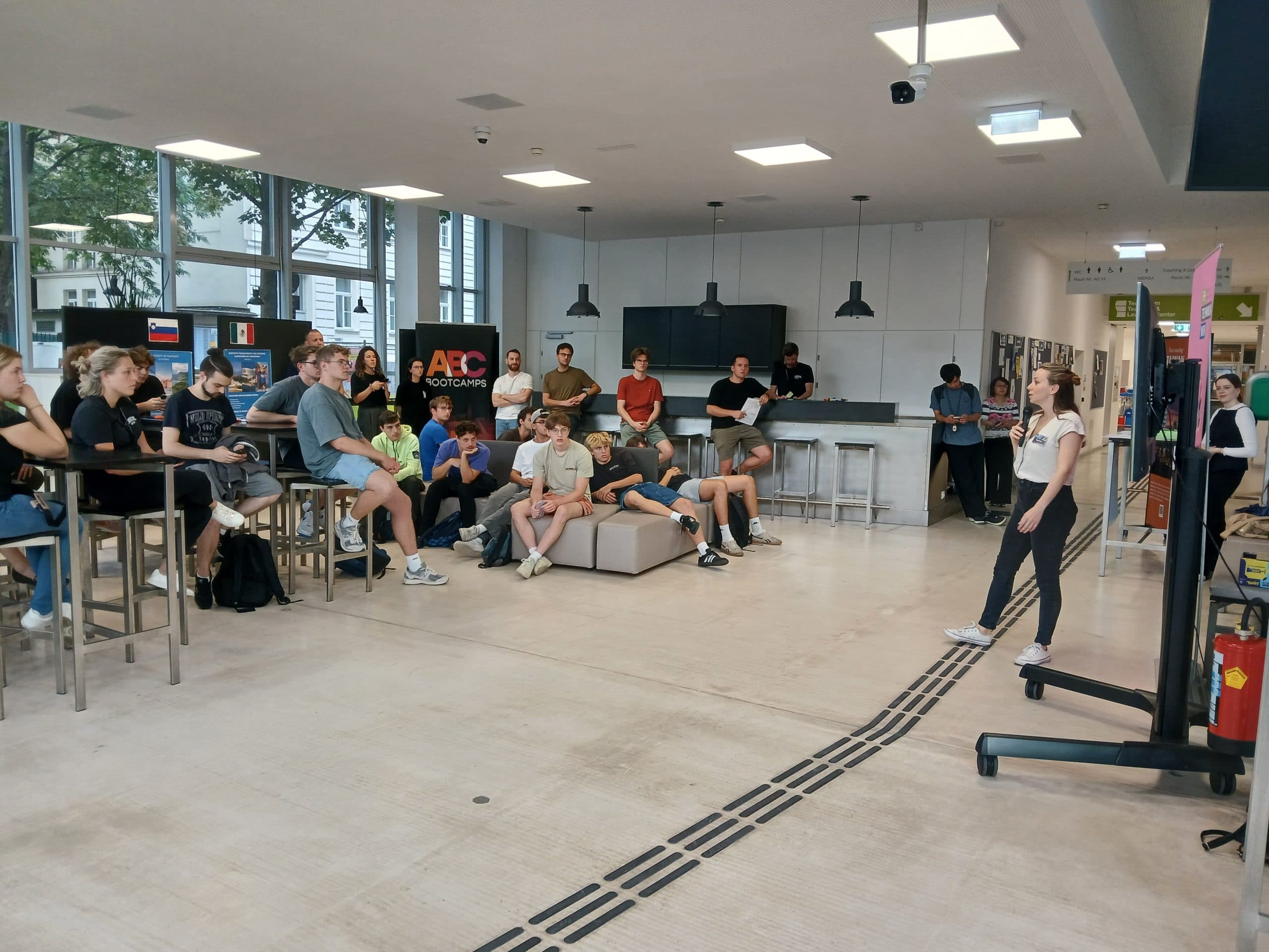 Eine Frau steht und spricht zu einer Gruppe junger Menschen, die in einem modernen, gut beleuchteten Klassenzimmer mit großen Fenstern und einem Küchenbereich im Hintergrund sitzen und aufmerksam zuhören.