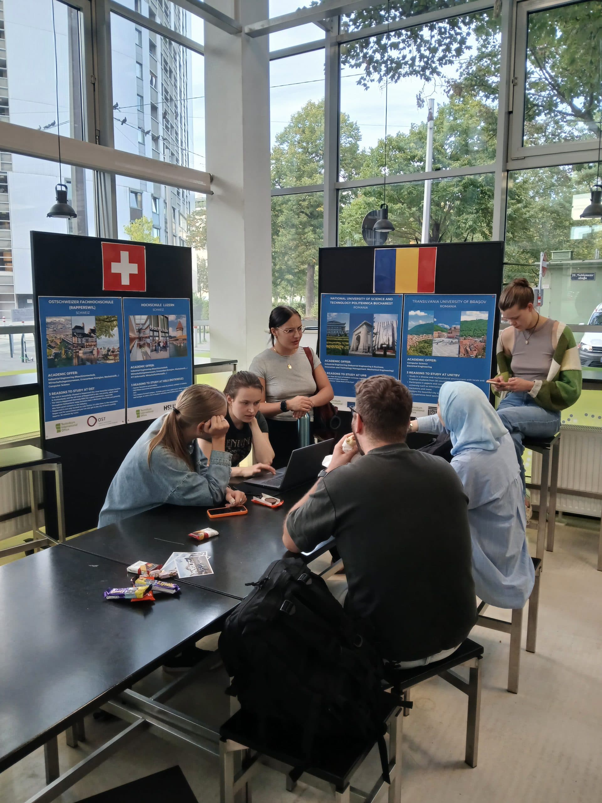 Eine Gruppe von sechs jungen Leuten sitzt und steht um einen Tisch mit Laptops und Snacks. Hinter ihnen hängen in einem hellen, verglasten Raum Plakatwände mit Bildern und Flaggen der Schweiz und Rumäniens.