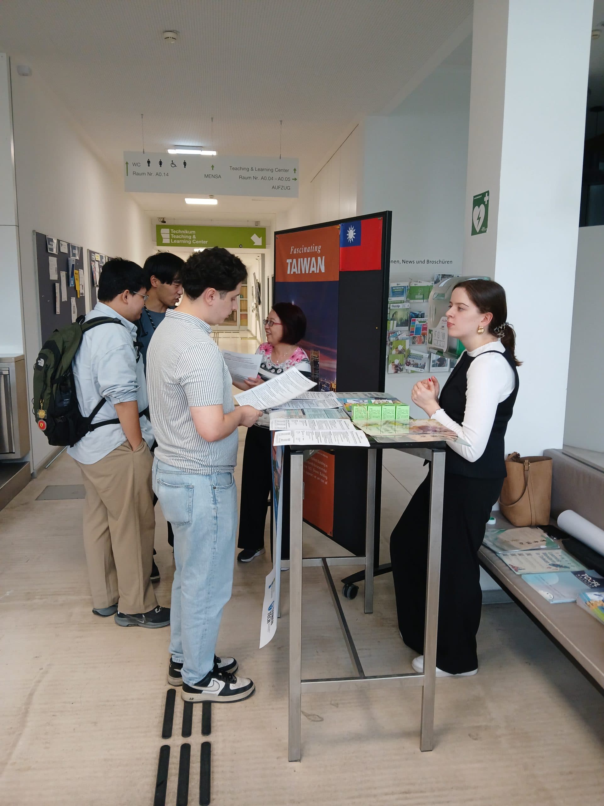 Eine Gruppe von Menschen steht an einem Informationsstand mit der Aufschrift "Fascinating Taiwan" und unterhält sich mit einer Frau hinter dem Tresen in einem modernen Flur, in dem Broschüren, Poster und Informationsmaterial ausliegen.