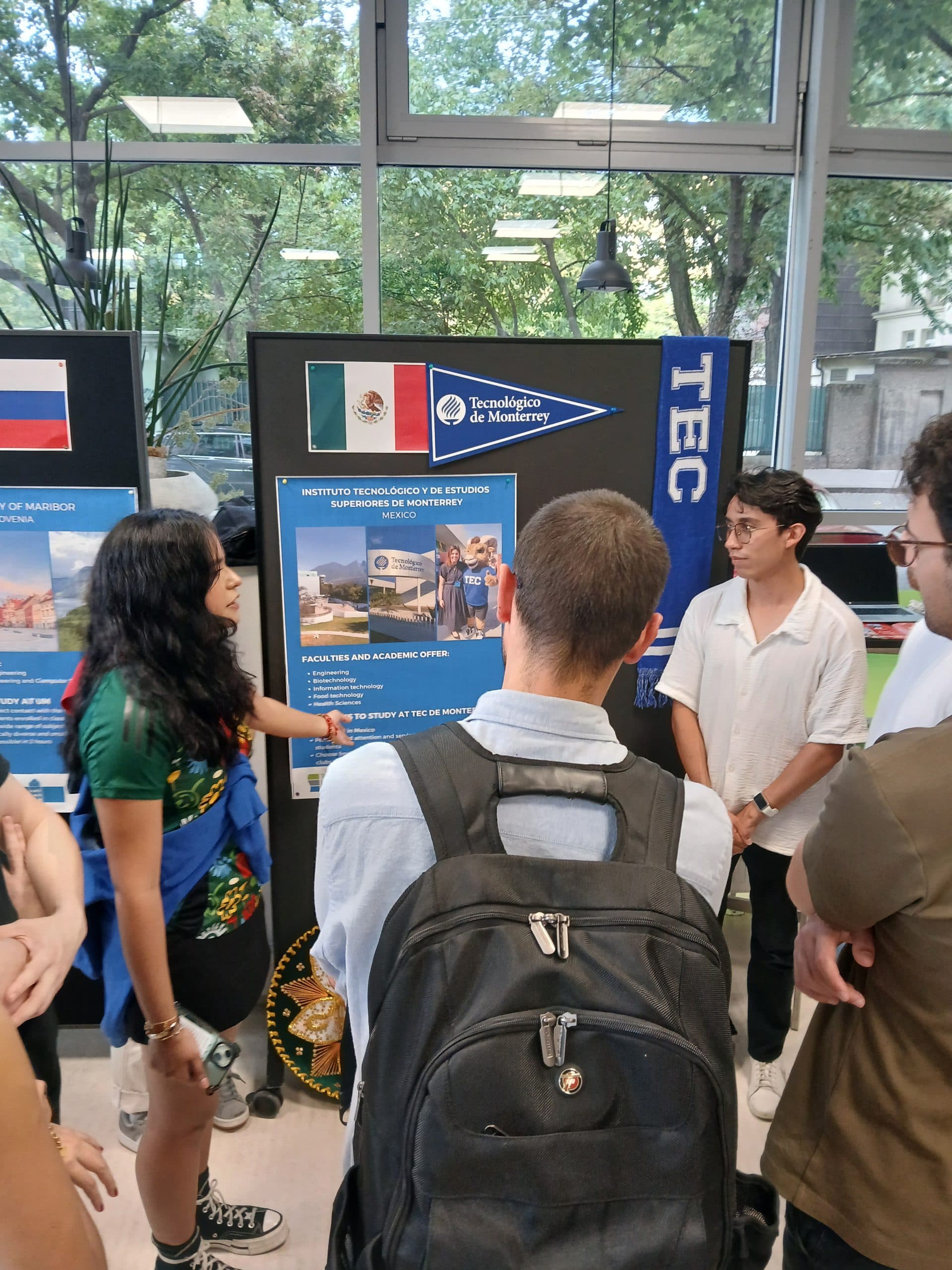Eine Frau und ein Mann präsentieren drei Personen, die sich um eine Schautafel mit Broschüren, Fahnen und Postern in einem Klassenzimmer versammelt haben, Informationen über Tecnológico de Monterrey.