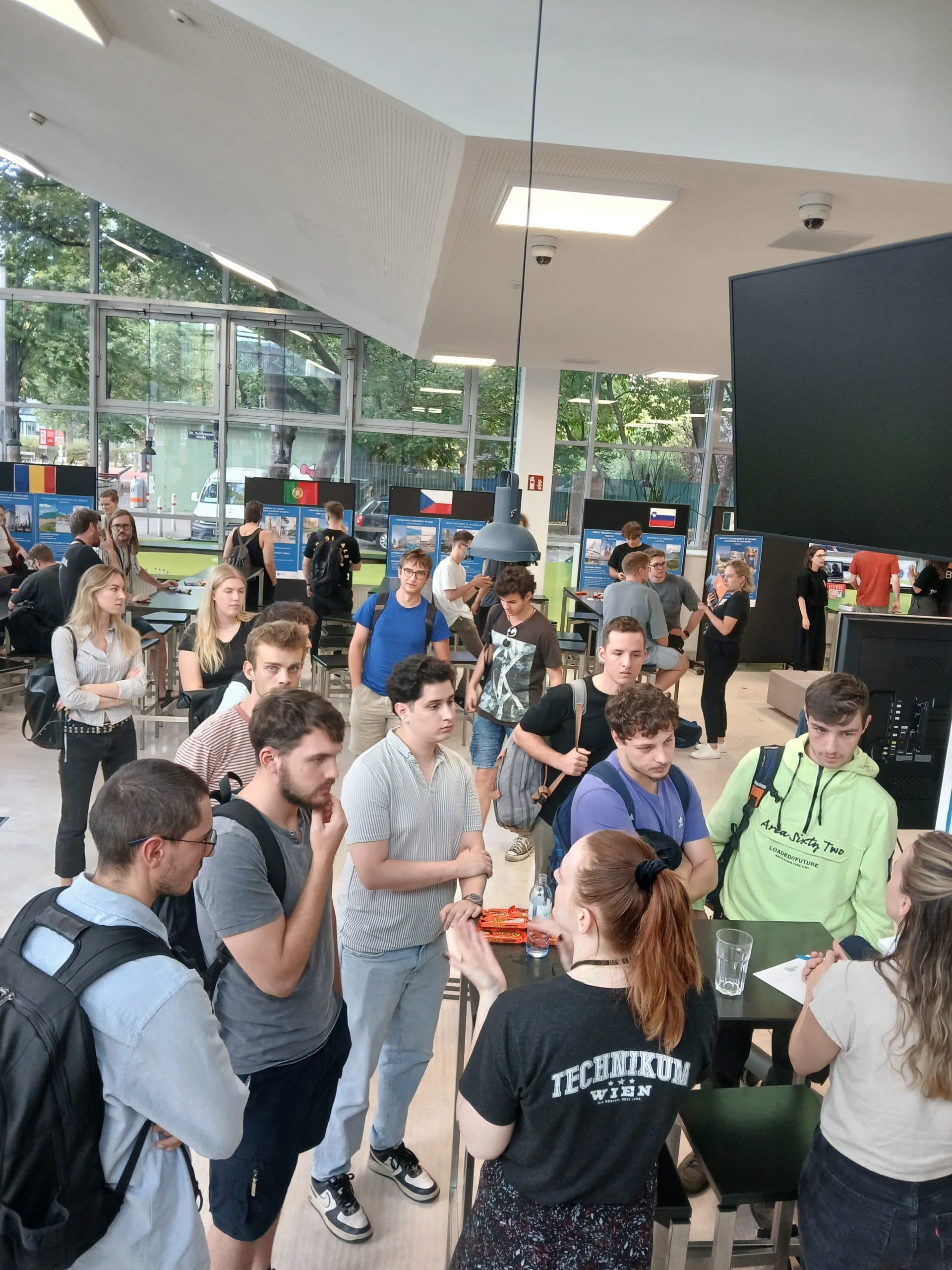 In einem hellen, modernen Raum mit großen Fenstern und Postern an den Wänden sitzt eine Gruppe von Studenten um einen Tisch herum. Die meisten hören aufmerksam einer Frau zu, die in der Mitte spricht.
