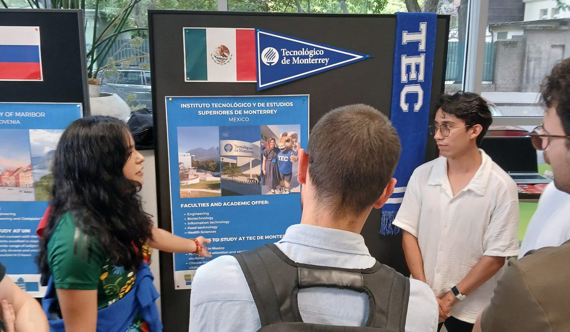 Eine Gruppe von Studenten steht um eine Schautafel des Tecnológico de Monterrey herum und hört einer Frau zu, die eine Rede hält; ein Student trägt einen Rucksack, ein anderer ein weißes Hemd. Im Hintergrund sieht man durch große Fenster Bäume.