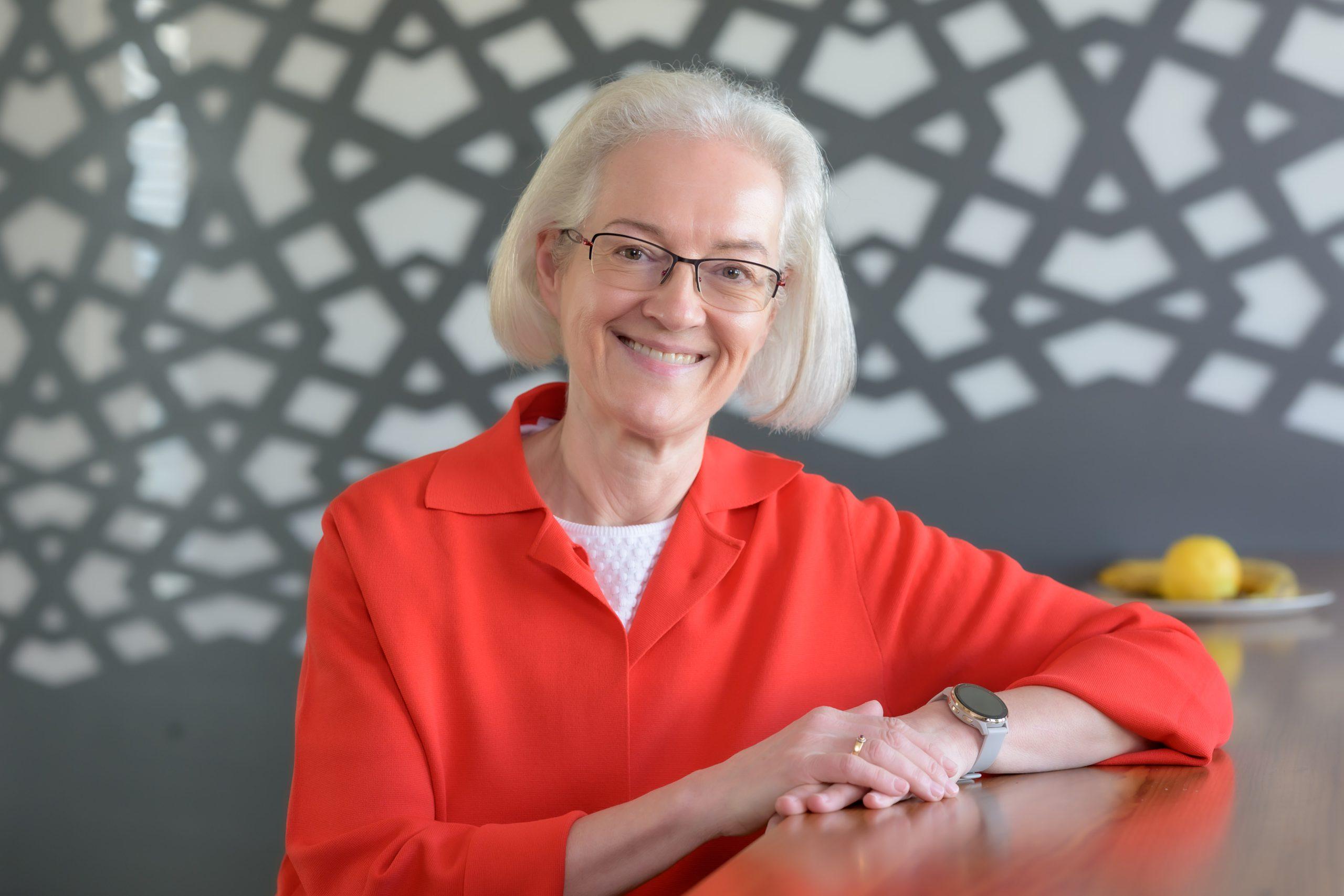 Eine ältere Frau mit weißem Haar und Brille, die ein leuchtend rotes Hemd trägt, sitzt lächelnd an einem Holztisch. Im Hintergrund sind eine geometrisch gemusterte Wand und ein Teller mit Zitronen zu sehen.