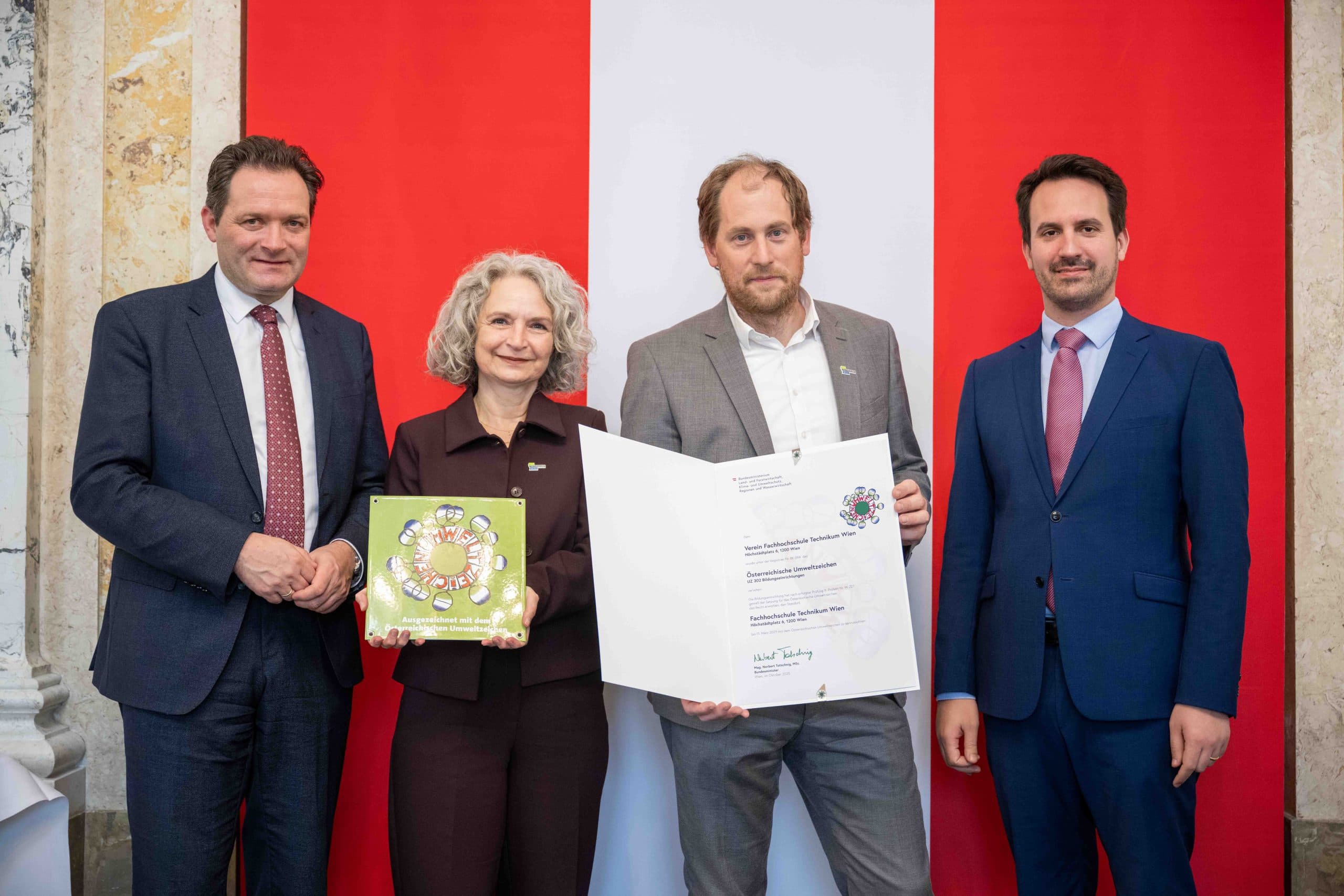 Vier formal gekleidete Personen stehen vor einem rot-weißen Hintergrund. Zwei von ihnen halten eine Urkunde und eine Auszeichnung in der Hand. Alle schauen in die Kamera und lächeln leicht.
