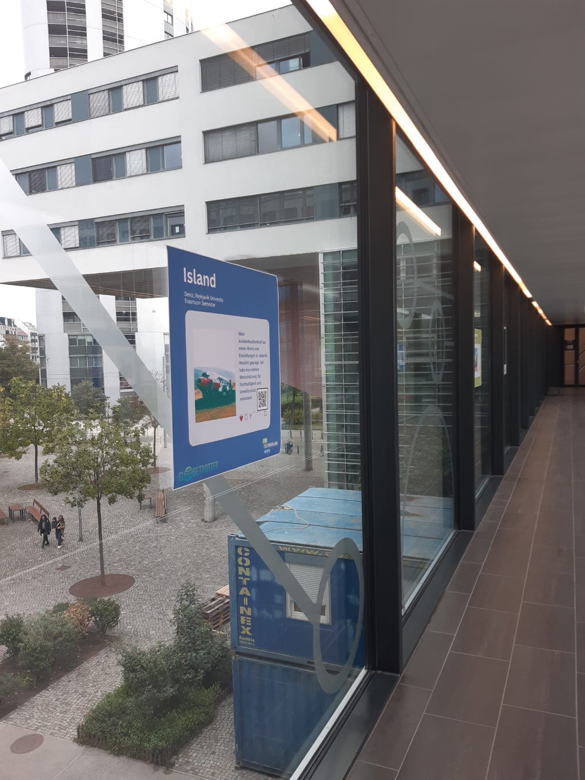 Ein großer Flur mit Fenstern blickt auf einen Hof mit Bäumen und modernen weißen Gebäuden. Am Fenster ist ein blaues Schild mit Informationen über Island angebracht, das vom Inneren des Flurs aus sichtbar ist.