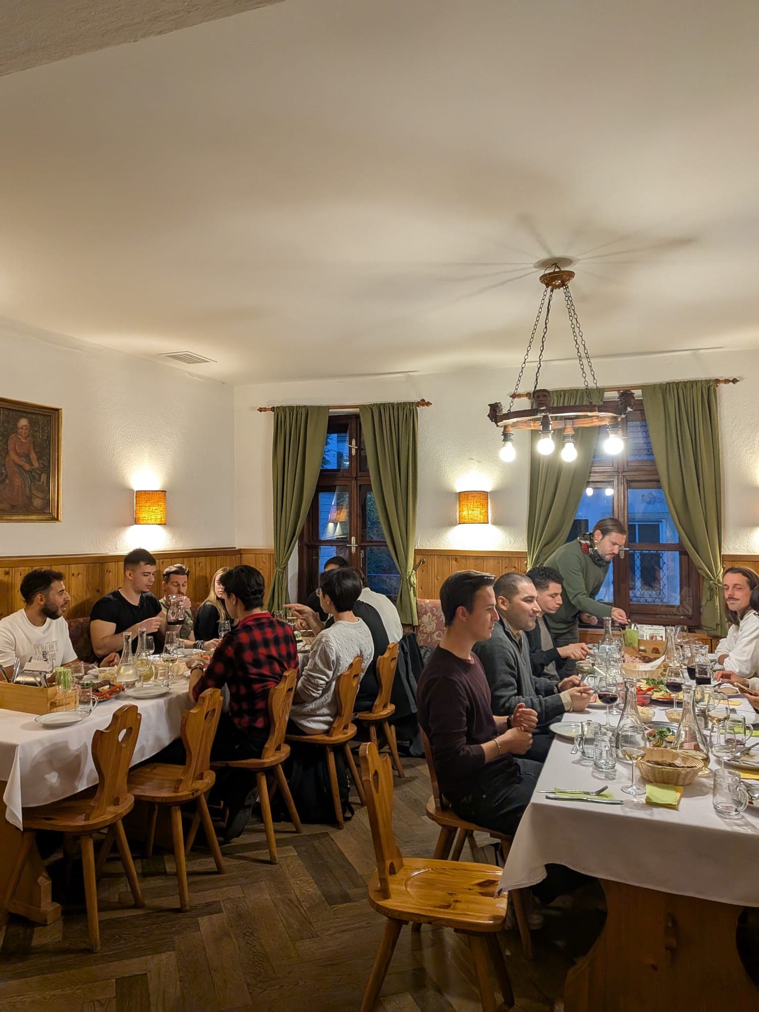 Eine Gruppe von Menschen sitzt an zwei langen Tischen in einem gemütlichen Restaurant, isst und unterhält sich. Der Raum ist mit Holzmöbeln, grünen Vorhängen und warmer Beleuchtung ausgestattet, was eine einladende Atmosphäre schafft. Speisen und Getränke sind auf den Tischen verteilt.
