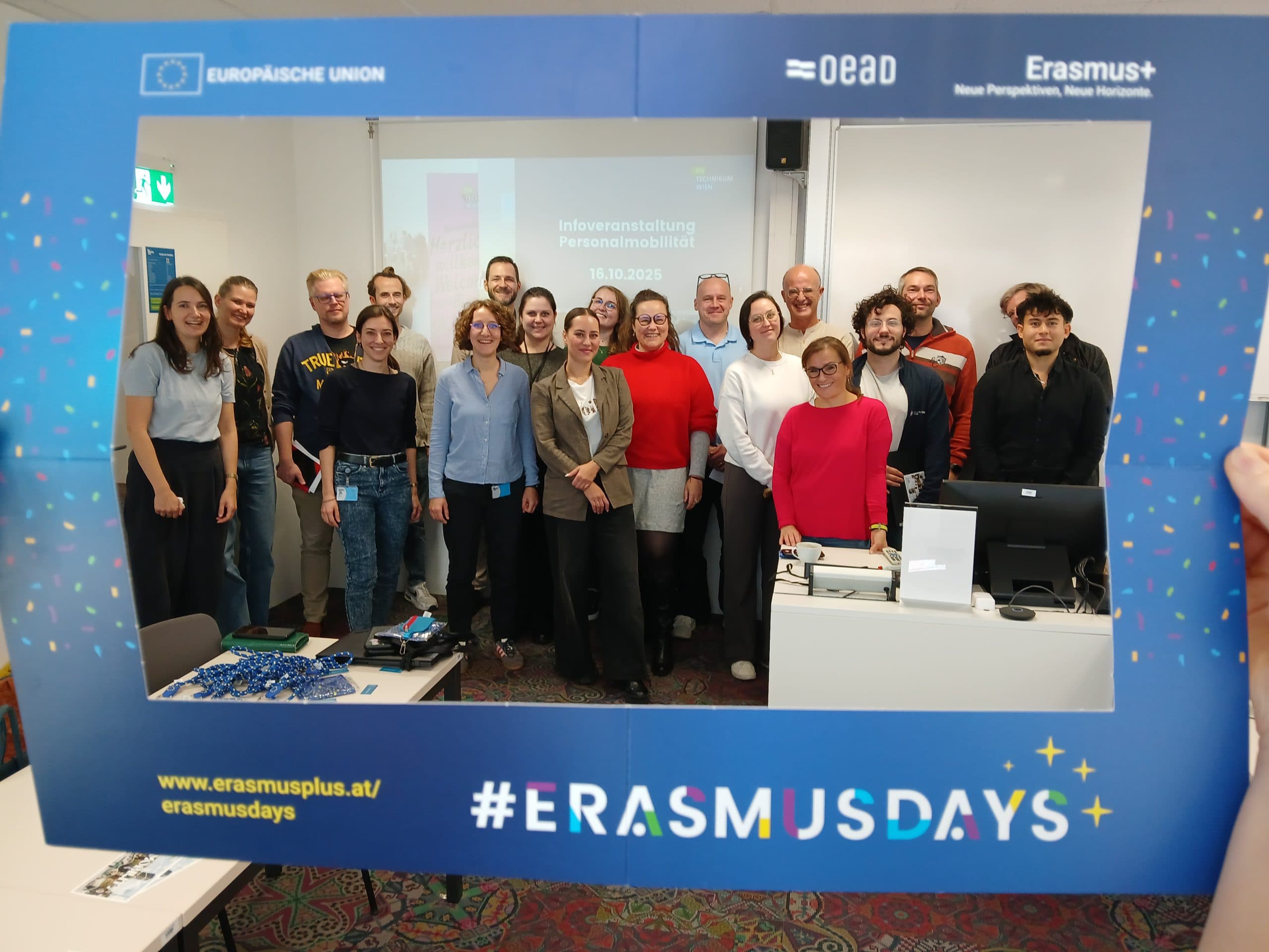 Eine Gruppe von lächelnden und posierenden Menschen in einem Klassenzimmer, eingerahmt von einem blauen #ErasmusDays-Rahmen. Im Hintergrund sind ein Bildschirm und Schreibtische mit Laptops zu sehen.