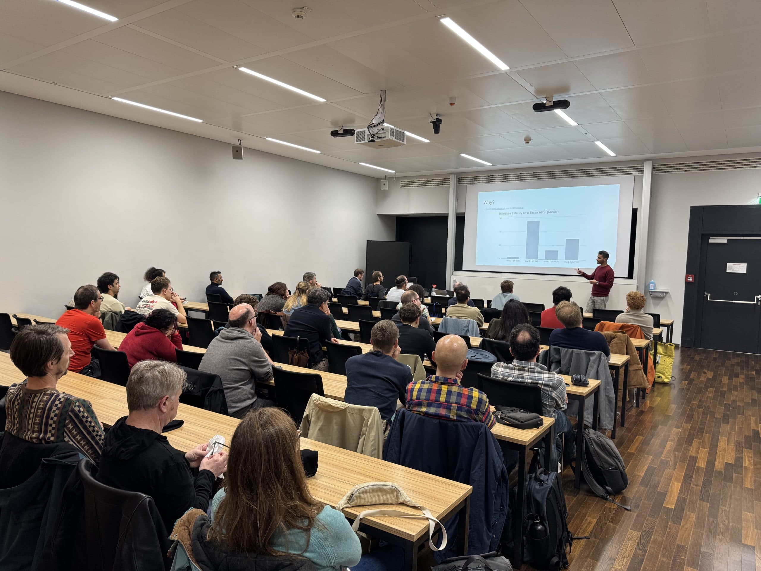 Eine Gruppe von Personen sitzt in einem Klassenzimmer und blickt auf einen Vortragenden, der vor einem Bildschirm steht, auf dem ein Balkendiagramm und Text angezeigt werden. Der Raum hat einen Holzboden, moderne Beleuchtung und Reihen von Holztischen und -stühlen.