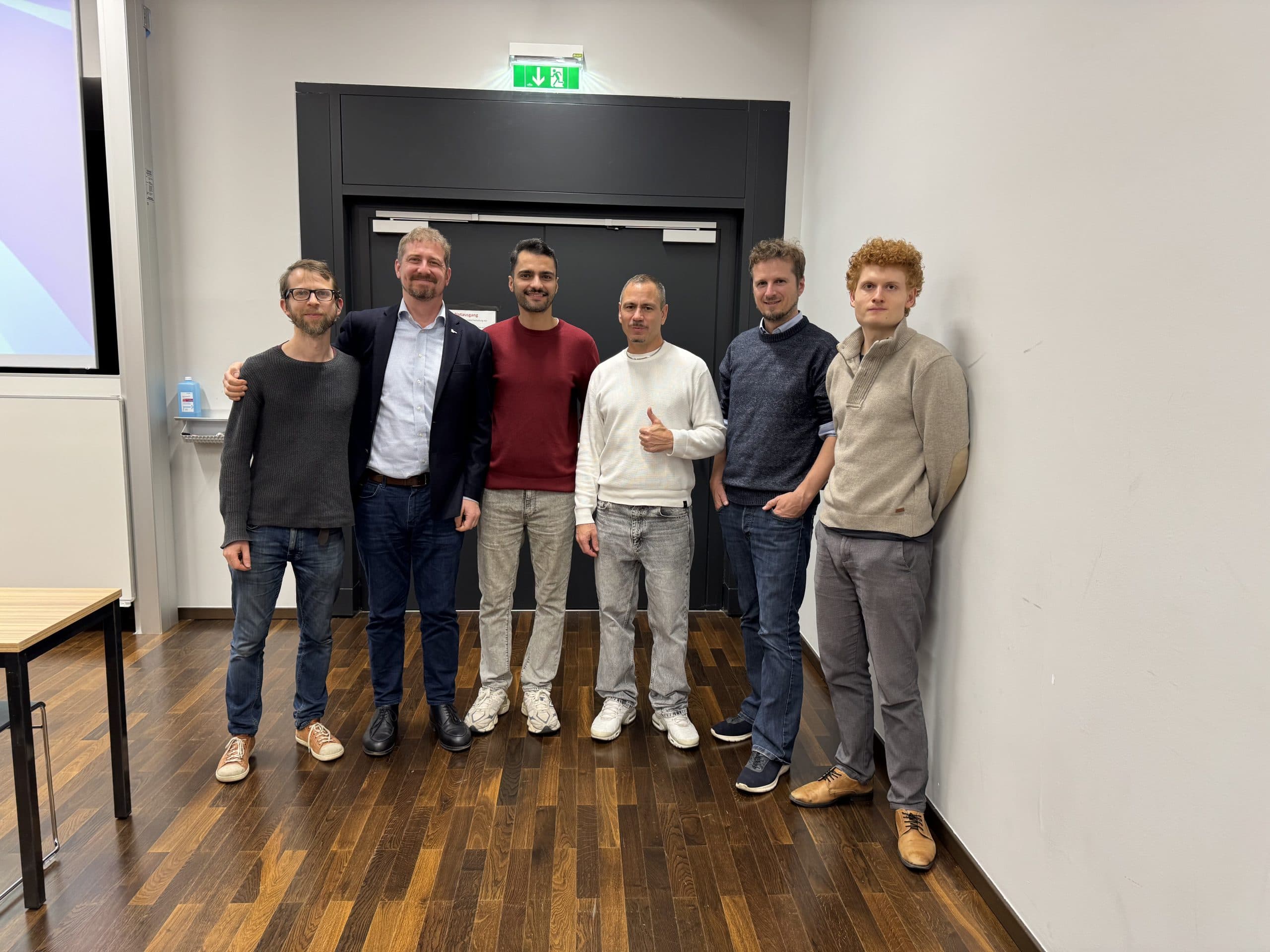Sechs Männer stehen nebeneinander in einem Raum mit Holzboden und weißen Wänden und posieren für ein Gruppenfoto. Ein Mann zeigt den Daumen nach oben. Hinter ihnen befinden sich eine schwarze Tür und ein grünes Ausgangsschild.