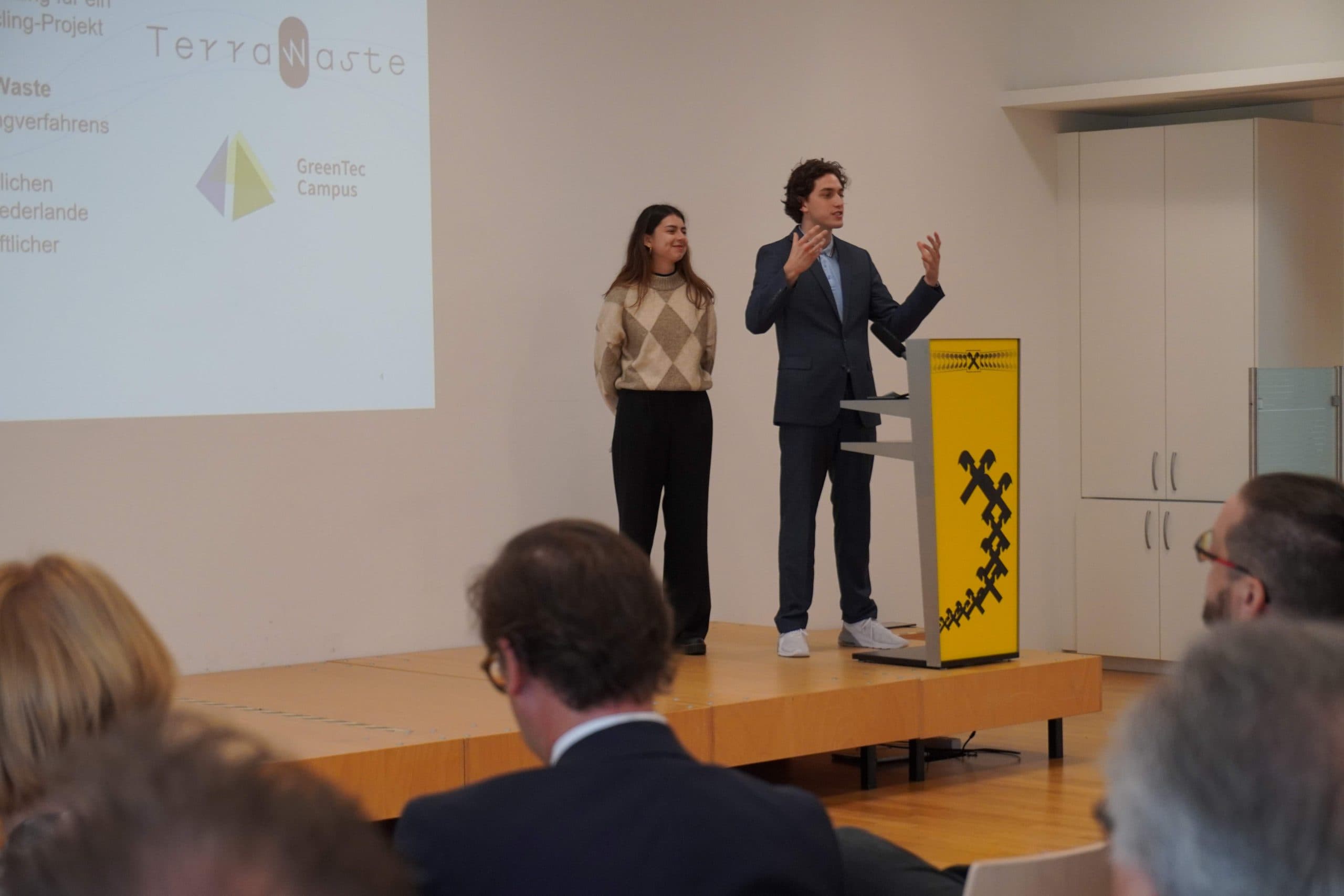 Zwei Personen stehen auf einer Bühne hinter einem gelben Podium und halten einen Vortrag vor dem Publikum. Eine Projektion an der Wand zeigt TerraWaste und Logos. Die Vortragenden scheinen enthusiastisch etwas zu erklären.