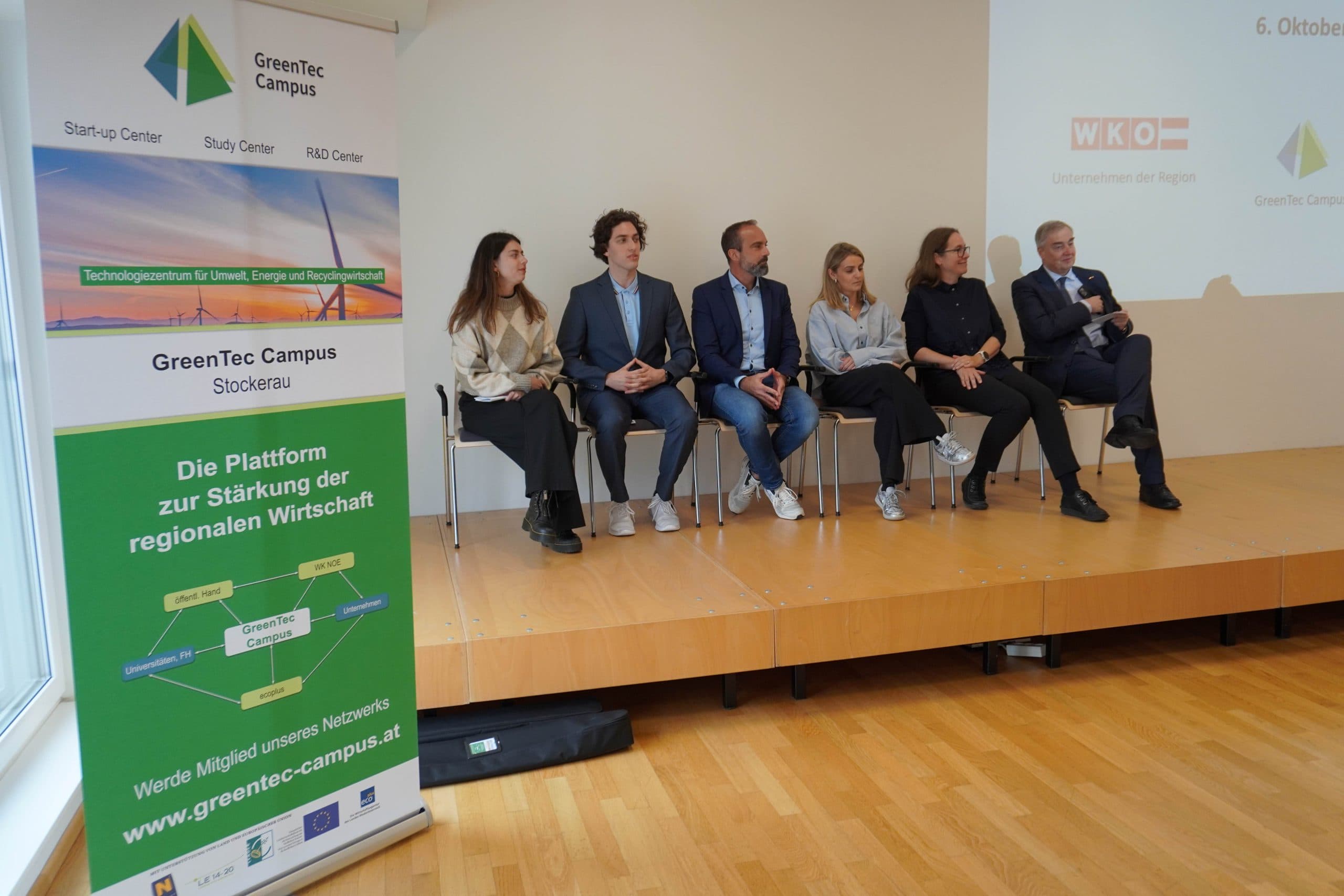 Sechs Personen sitzen auf einer Bühne vor einem leeren Bildschirm und scheinen an einer Podiumsdiskussion teilzunehmen. Auf der linken Seite des Bildes steht ein GreenTec Campus-Banner. Die Kulisse ist ein moderner, heller Raum mit Holzfußboden.