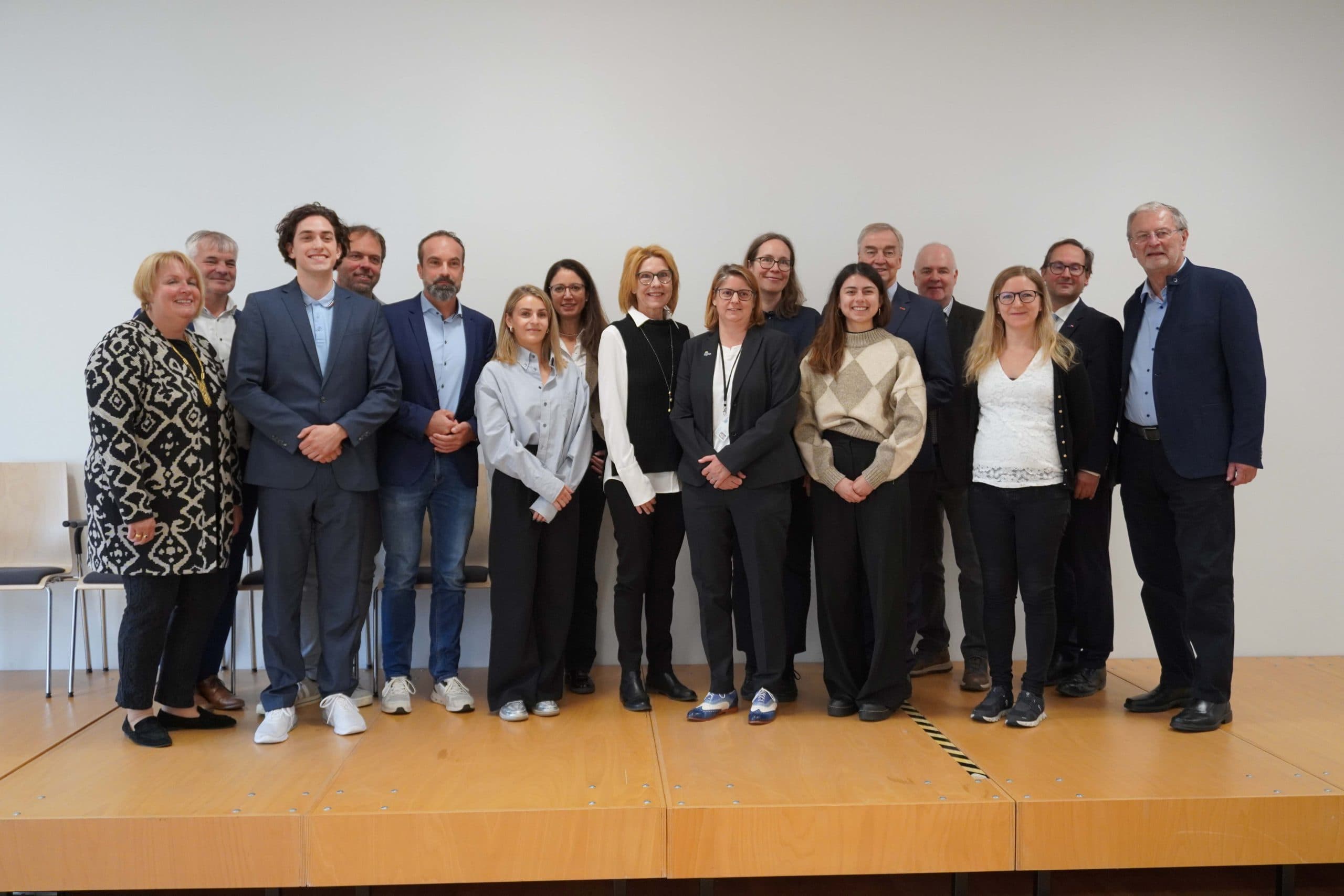 Eine Gruppe von fünfzehn Personen steht auf einer hölzernen Bühne zusammen und posiert lächelnd für ein Gruppenfoto vor einer schlichten weißen Wand. Die meisten von ihnen tragen Geschäftskleidung oder elegante Freizeitkleidung.