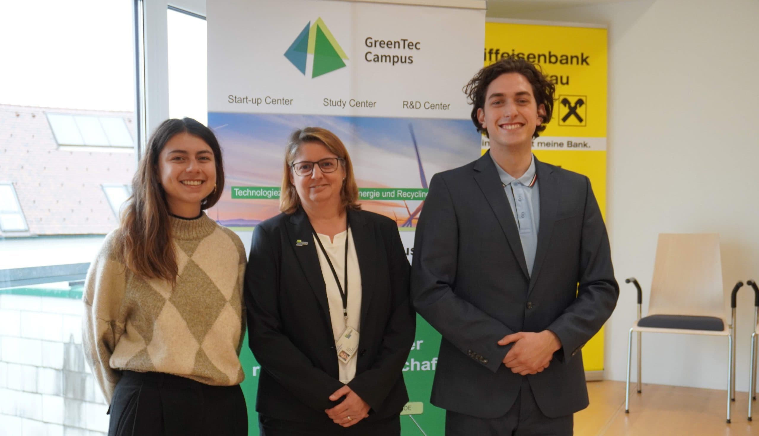 Drei Personen stehen lächelnd in einem Raum vor einem GreenTec Campus-Banner und einem gelben Raiffeisenbank-Schild. Links stehen zwei Frauen, rechts ein Mann im Anzug. Im Hintergrund sind ein Fenster und Stühle zu sehen.