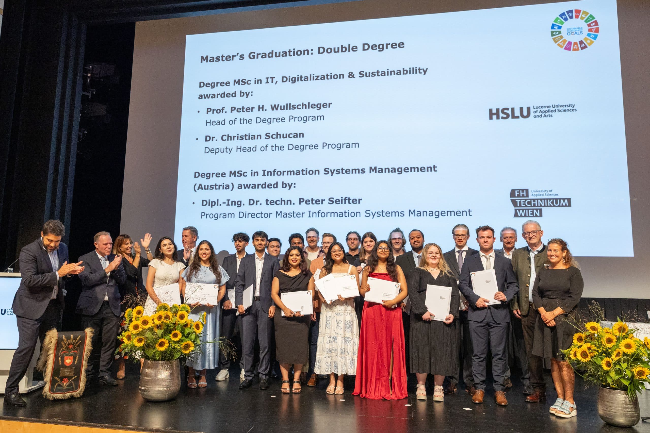Eine Gruppe von Absolventen und Fakultätsmitgliedern steht auf der Bühne, lächelnd und mit ihren Diplomen in der Hand, bei einer Master-Abschlussfeier. Sonnenblumen schmücken die Bühne, und auf der Leinwand hinter ihnen wird ein Glückwunschdia gezeigt.