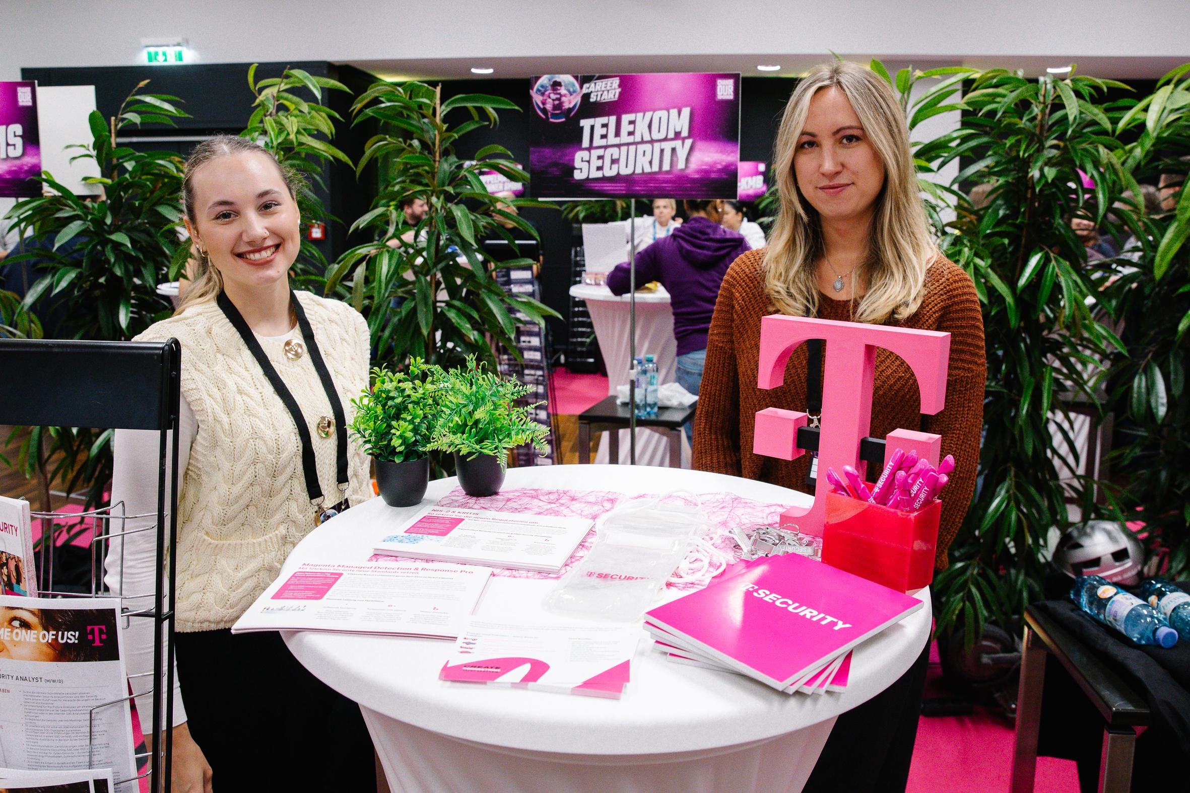 Zwei Frauen stehen hinter einem runden Tisch mit Telekom Security-Materialien, darunter Broschüren, Stifte und ein großes rosafarbenes "T"-Logo, an einem mit Pflanzen und hellen Bannern geschmückten Stand in einem modernen Ambiente.