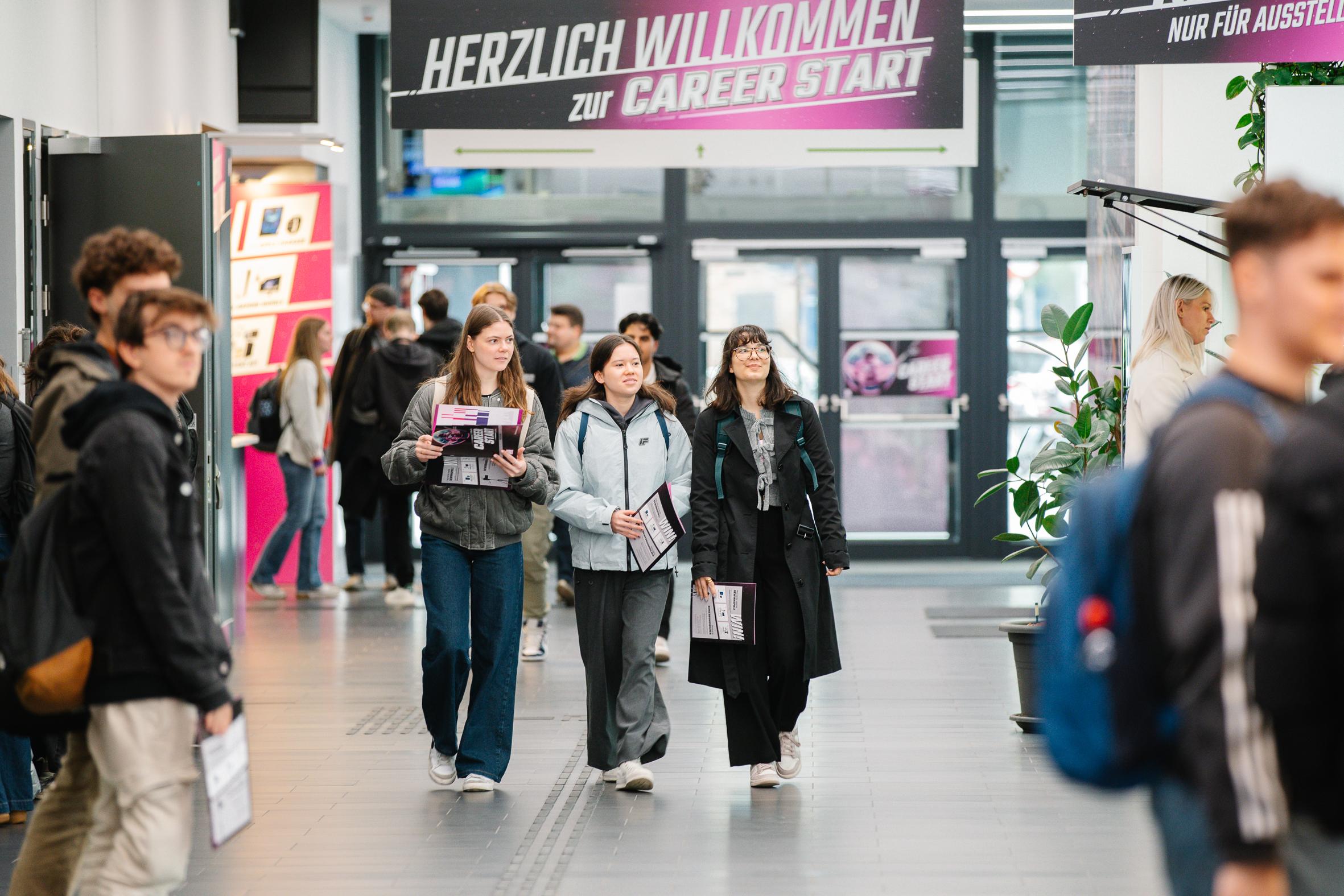 Eine Gruppe junger Leute geht mit Broschüren in der Hand durch eine helle Messehalle. Auf den Bannern darüber steht "Herzlich willkommen zum Career Start". Im Hintergrund sind andere Teilnehmer und Messestände zu sehen.