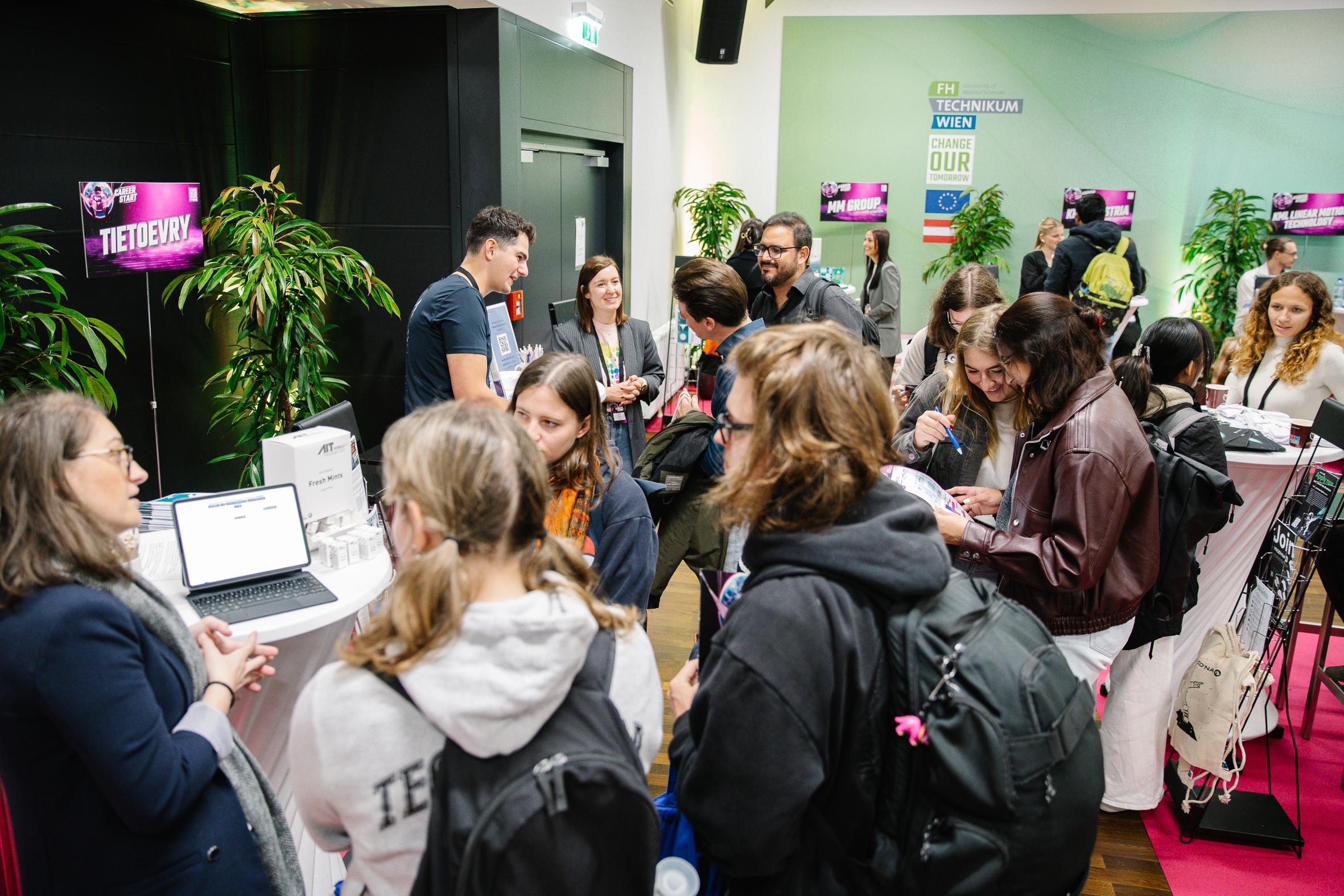 Eine Gruppe von Menschen versammelt sich bei einer Indoor-Veranstaltung um einen Stand und beschäftigt sich mit den Vortragenden und den technischen Displays. Einige schauen auf Laptops, während andere sich unterhalten oder Notizen machen. Die Umgebung ist modern mit Pflanzen und Beschilderung.