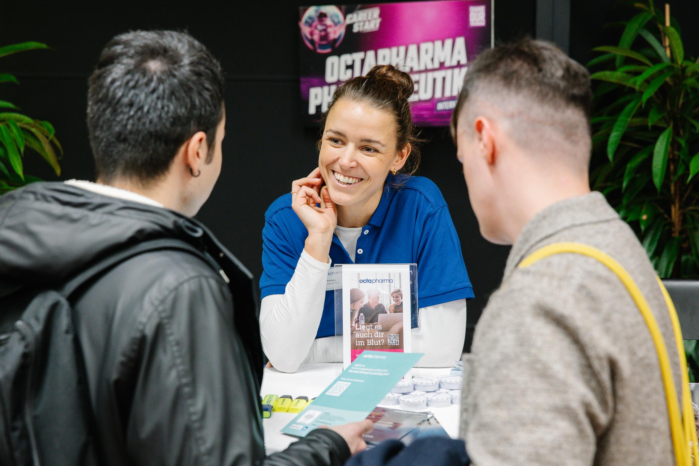 Eine Frau in einem blauen Hemd lächelt und spricht mit zwei Personen an einem Stand. Auf dem Tisch liegen Broschüren und Werbematerialien, und auf einem Plakat hinter ihr steht OCTAPHARMA PHARMA SOLUTIONS.