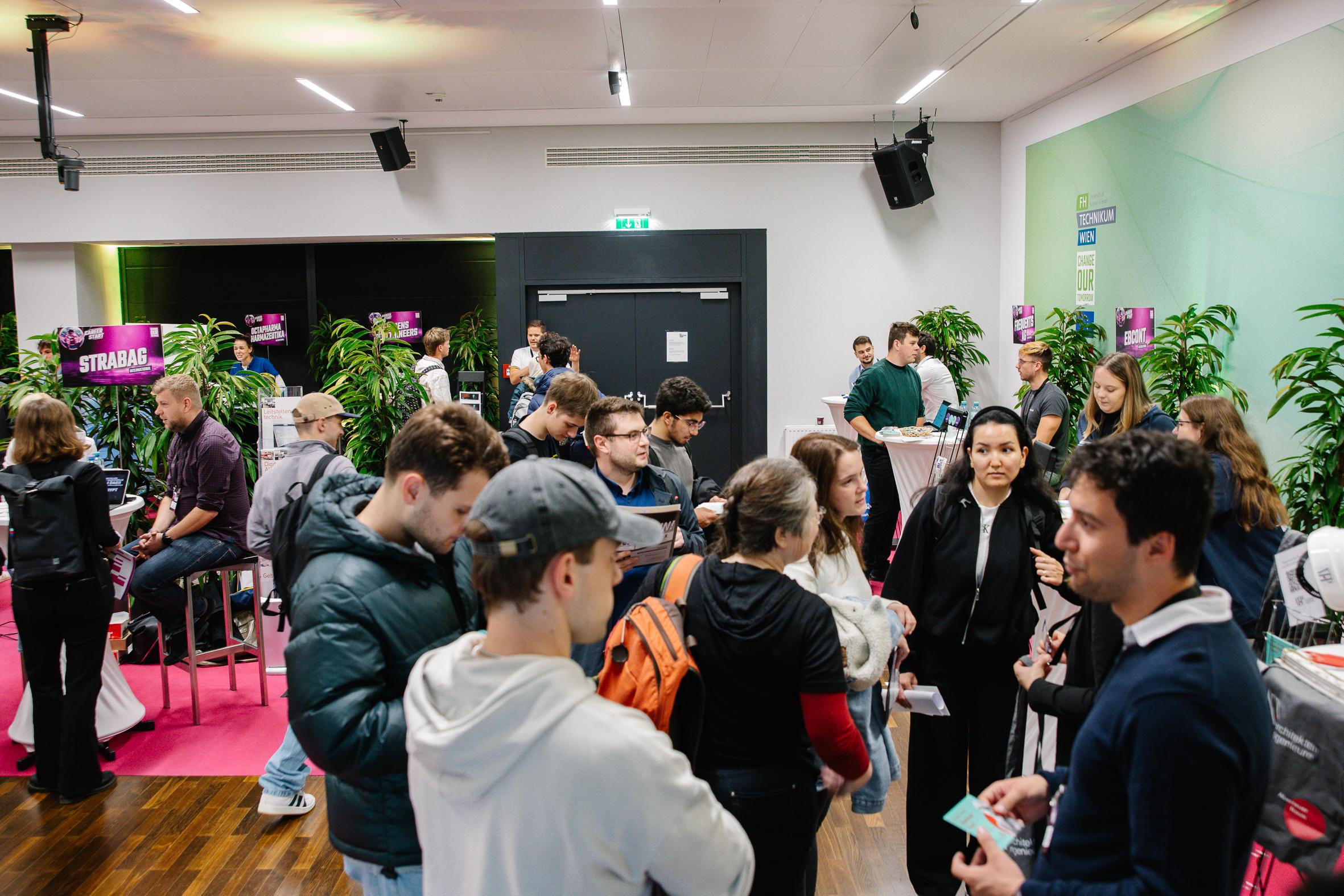Eine große Gruppe von Menschen interagiert auf einer Indoor-Veranstaltung oder -Messe, wobei einige in der Schlange stehen und andere sich in kleinen Gruppen unterhalten. Grüne Pflanzen und bunte Schilder schmücken den modernen Raum.