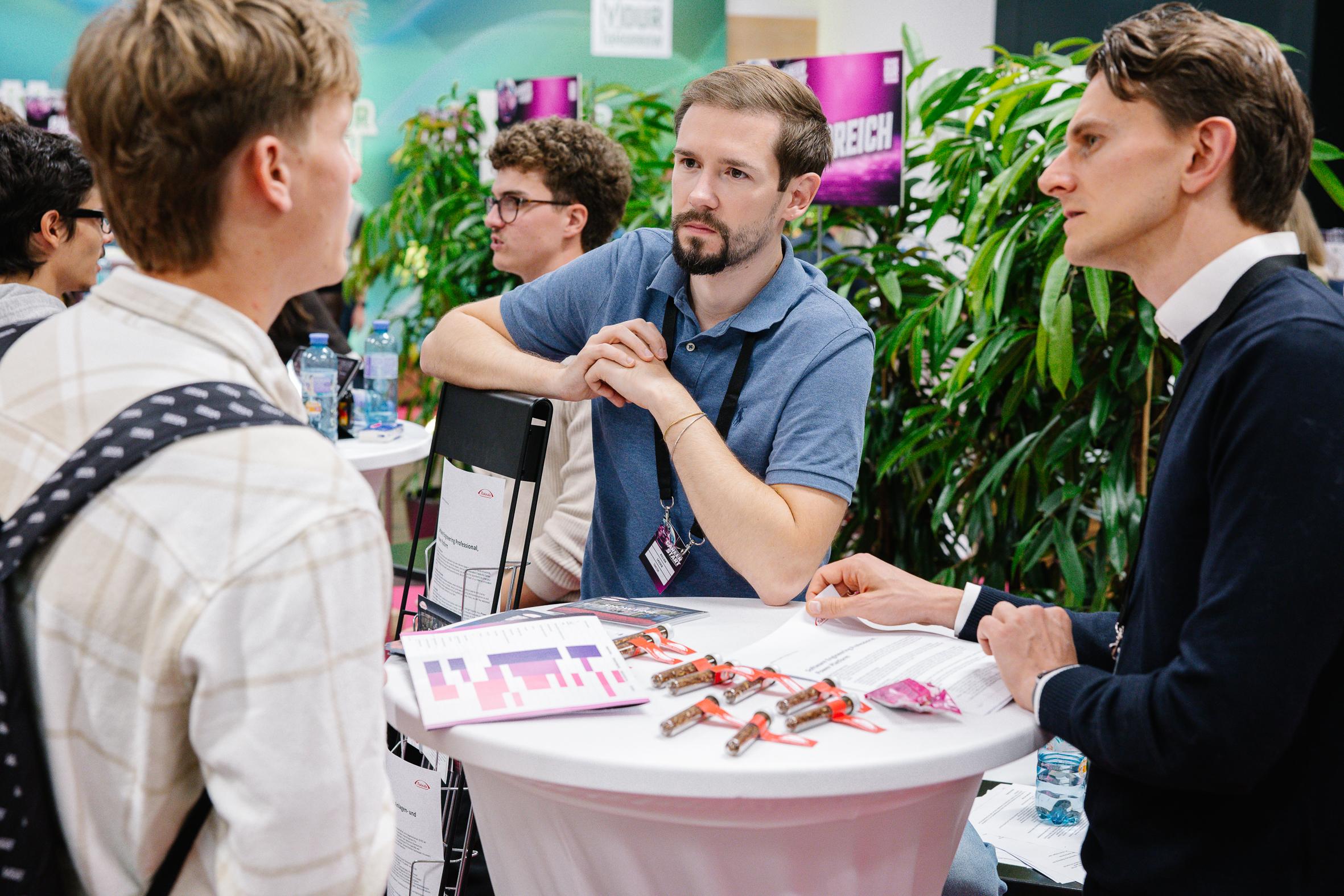 Drei Männer führen ein konzentriertes Gespräch an einem kleinen runden Tisch, der mit Papieren, Schokoriegeln und Stiften bedeckt ist. Zwei Männer sitzen, einer steht, und im Hintergrund stehen Pflanzen und digitale Bildschirme, die auf eine professionelle Veranstaltung hindeuten.