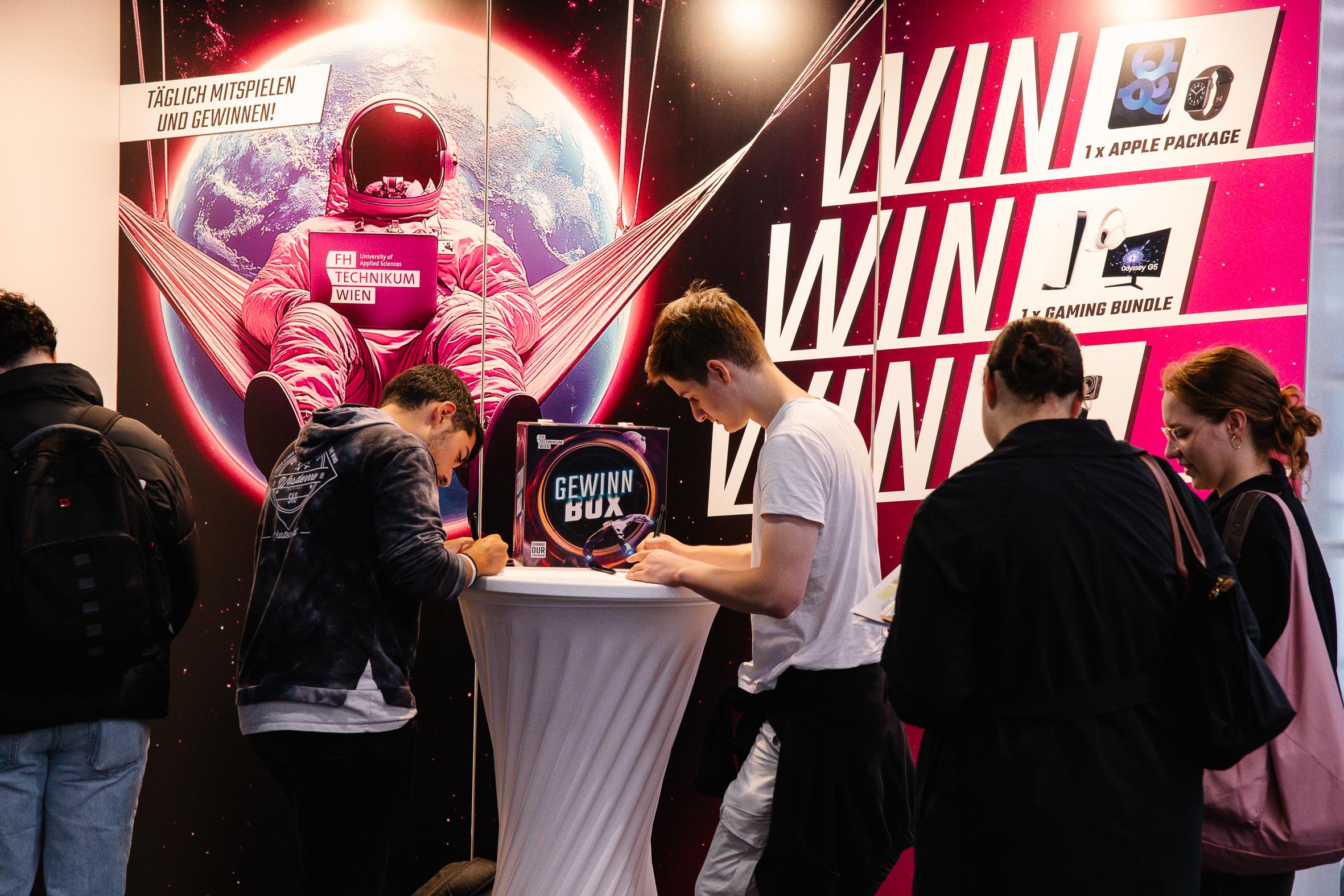 Mehrere Jugendliche stehen an einem hohen runden Tisch und schreiben Formulare vor einem farbenfrohen Stand mit einem rosafarbenen Astronauten und einem großen "WIN WIN WIN"-Schriftzug, der für einen Wettbewerb mit Preisen wie einem Apple-Paket und einem Spielepaket wirbt.