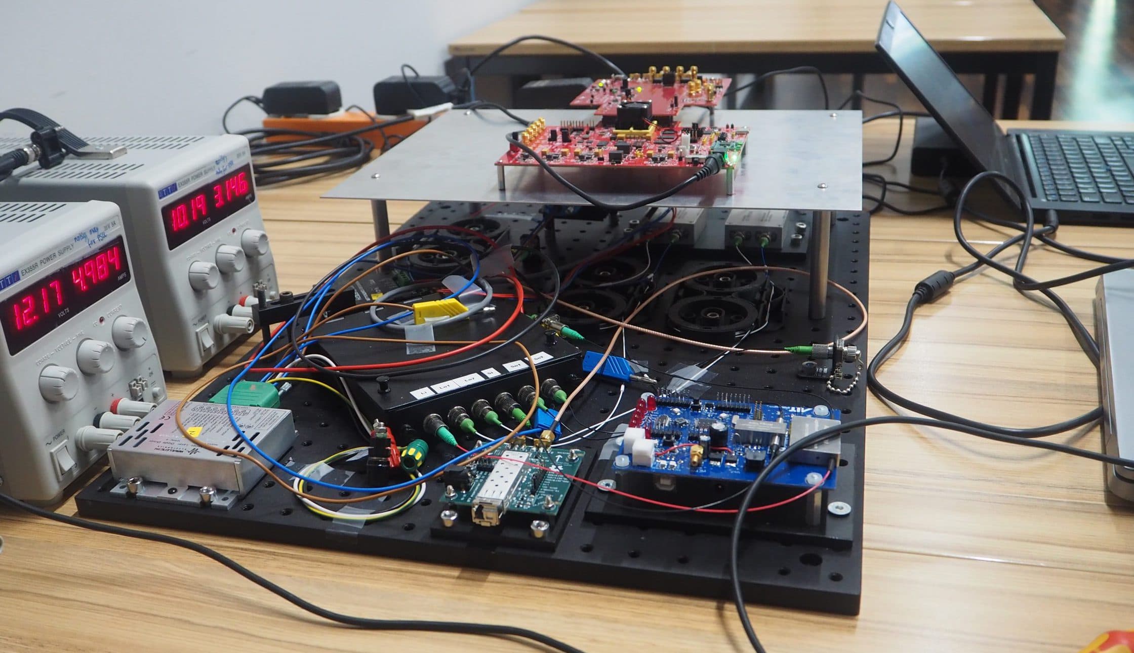 Ein elektronischer Arbeitsplatz mit Leiterplatten, Netzteilen, die Spannung und Stromstärke anzeigen, verschiedenen Kabeln und einem Laptop auf einem Holztisch. Der Aufbau scheint Teil eines Experiments oder eines Testverfahrens zu sein.