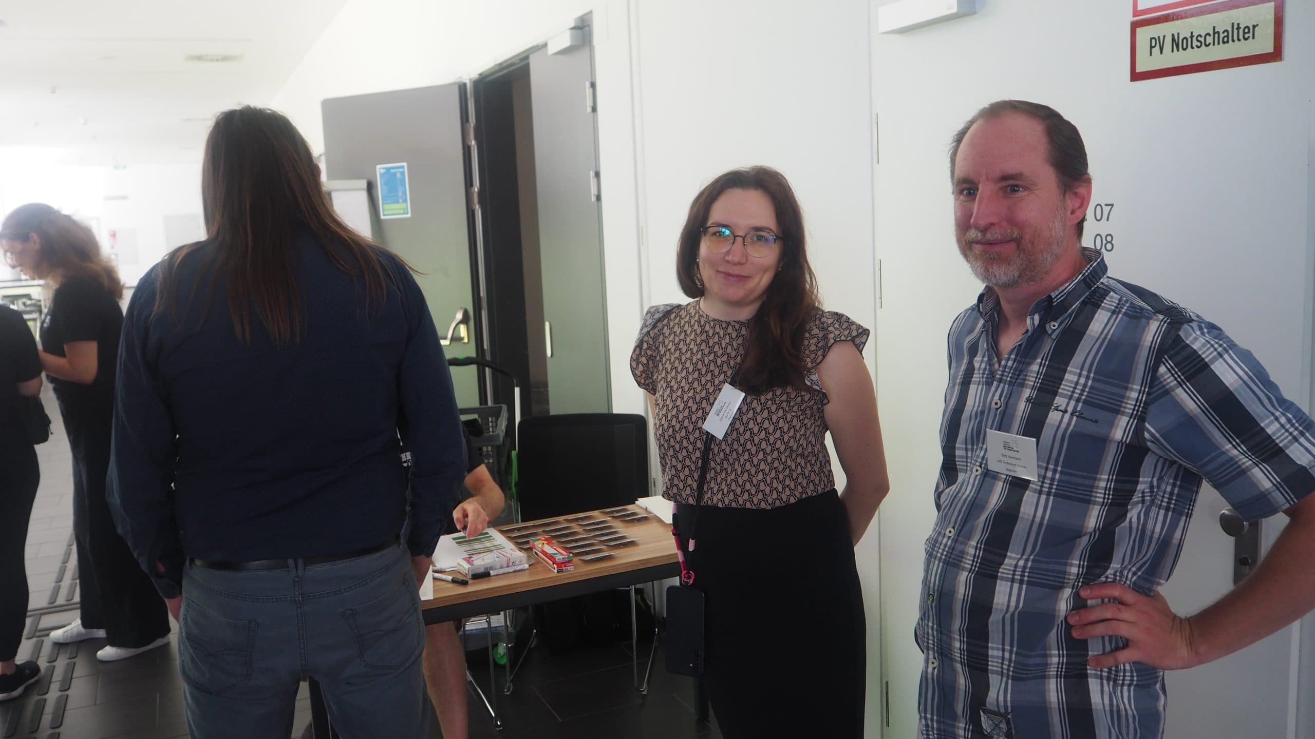 Zwei Personen stehen lächelnd in der Nähe eines Eingangs bei einer Veranstaltung in einem Gebäude, wobei im Hintergrund ein Tisch und andere Teilnehmer zu sehen sind. Die Frau trägt eine Brille und eine gemusterte Bluse; der Mann trägt ein kariertes Hemd und ein Namensschild.