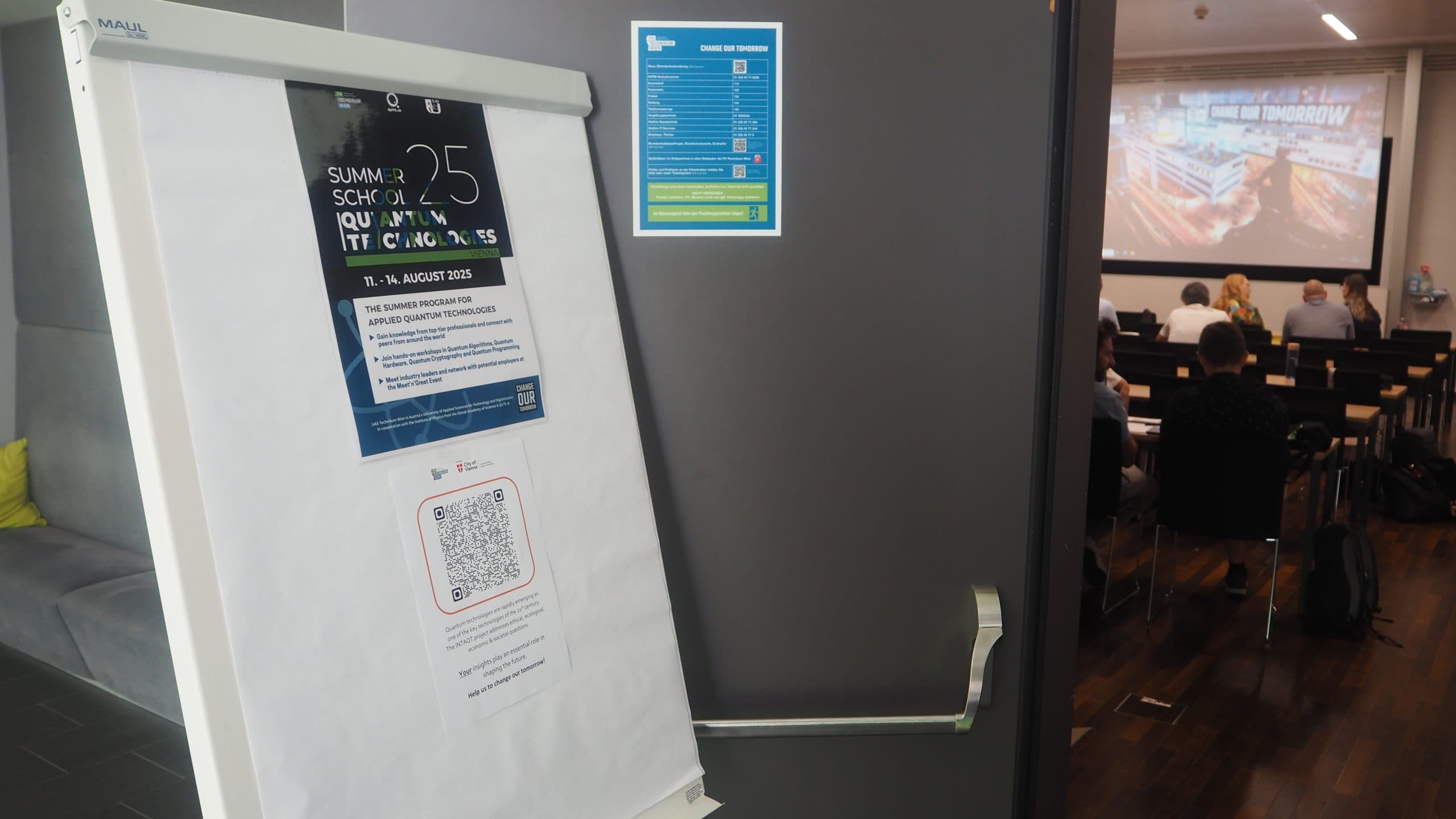 Ein Flipchart mit einem Poster und einem QR-Code steht neben der Tür zu einem Konferenzraum, in dem die Teilnehmer vor einer Projektionsfläche sitzen, auf der eine Präsentation gezeigt wird. Ein blaues Informationsblatt hängt an der Wand neben der Tür.