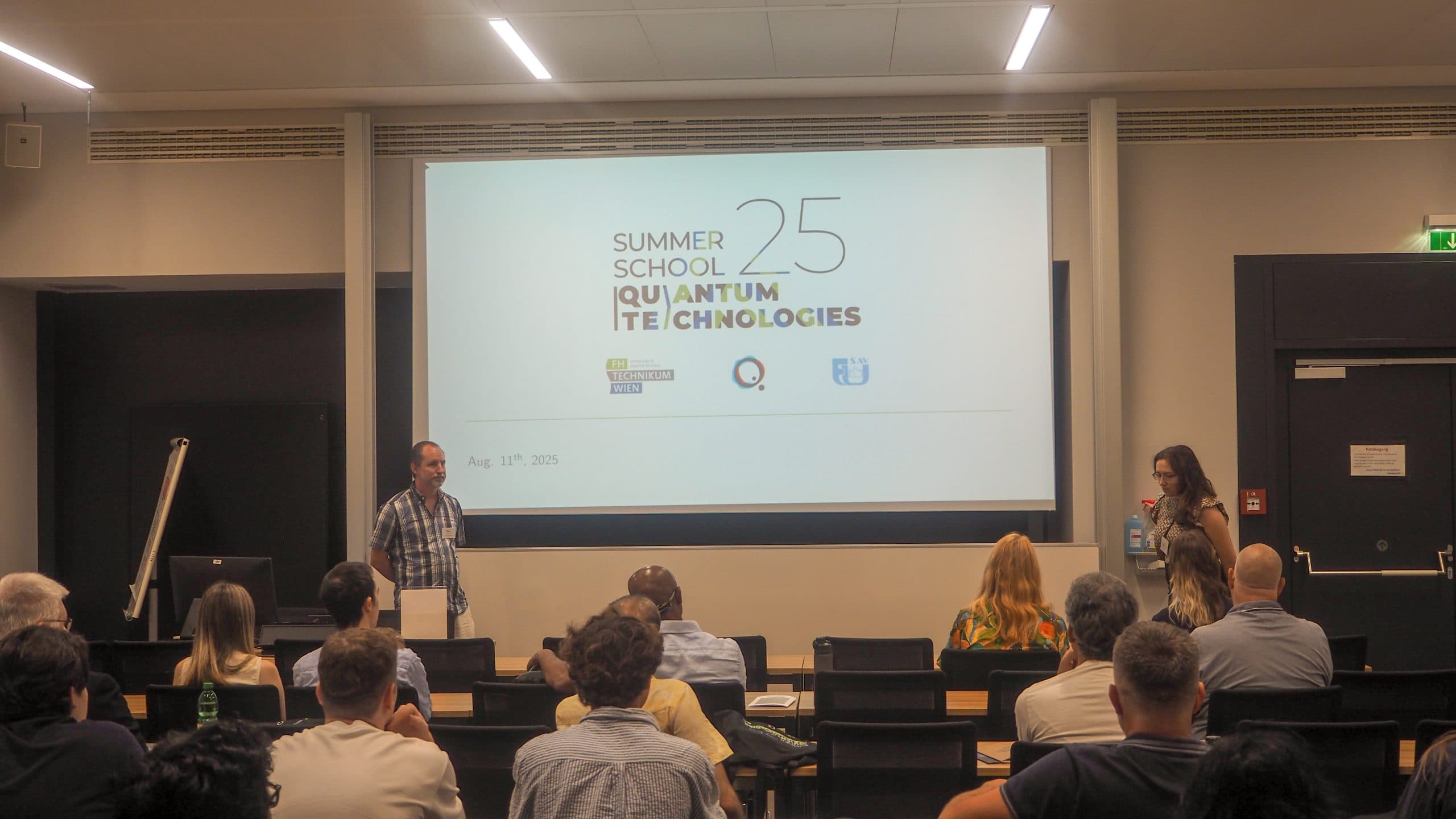 Ein Klassenzimmer mit Personen, die vor einem Bildschirm mit der Aufschrift Summer School 25 Quantum Technologies sitzen. Zwei Vortragende stehen vorne, einer an einem Podium, der andere hält ein Mikrofon.