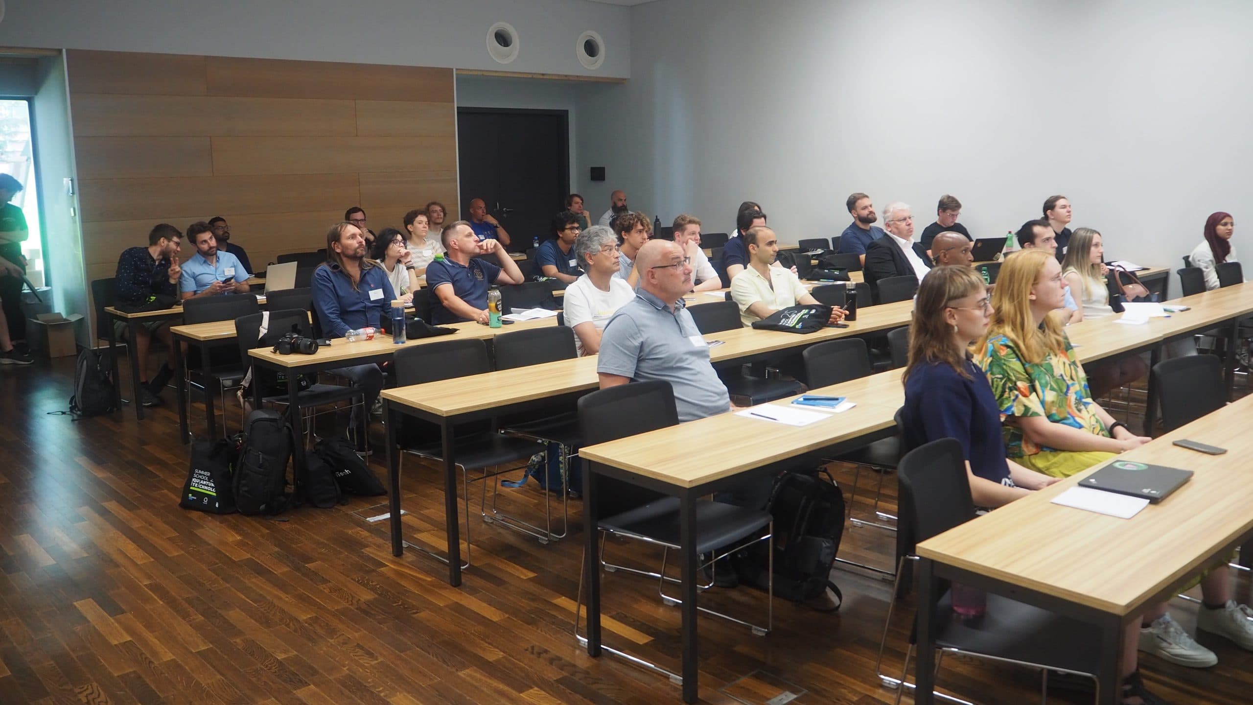 Eine Gruppe von Personen sitzt an Tischen in einem modernen Klassenzimmer oder Konferenzraum und blickt aufmerksam nach vorne. Der Raum hat Holzböden, helle Wände und natürliches Licht durch ein Fenster auf der linken Seite.