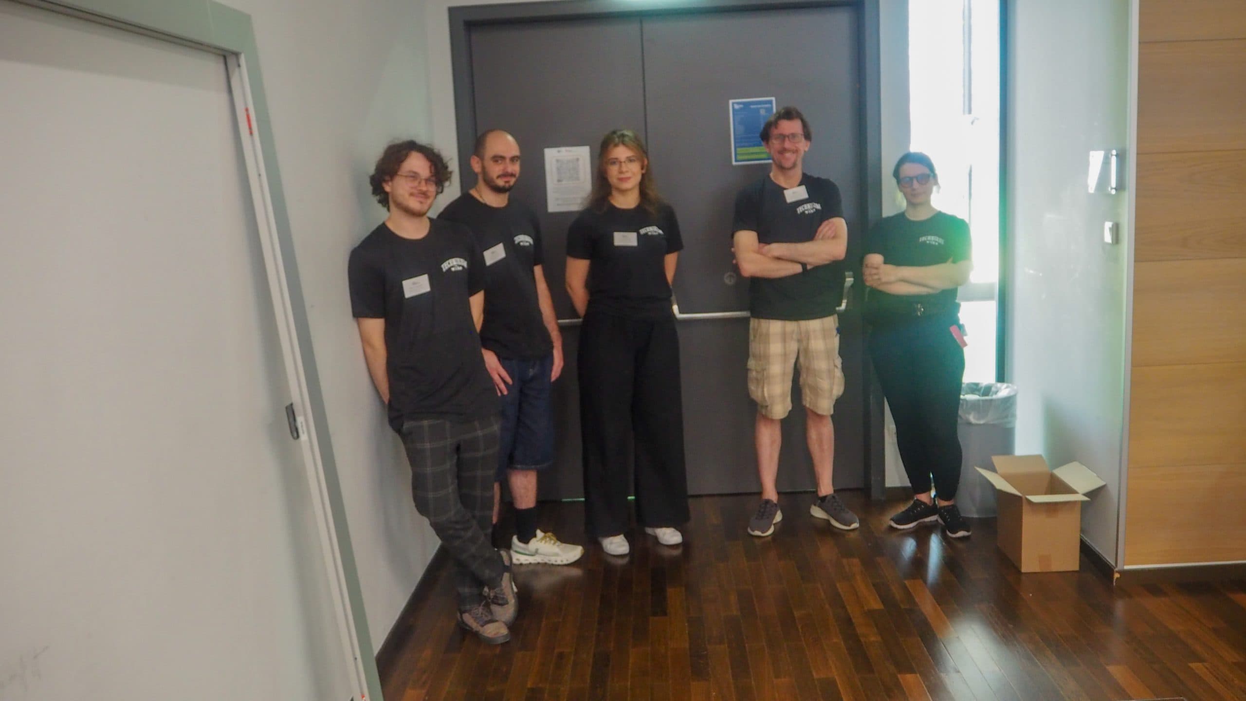 Fünf Personen stehen in einem Innenraum in einer Reihe, alle tragen passende schwarze T-Shirts. Sie lächeln in die Kamera. Der Boden ist aus Holz, und in der Nähe stehen ein Karton und ein Mülleimer.