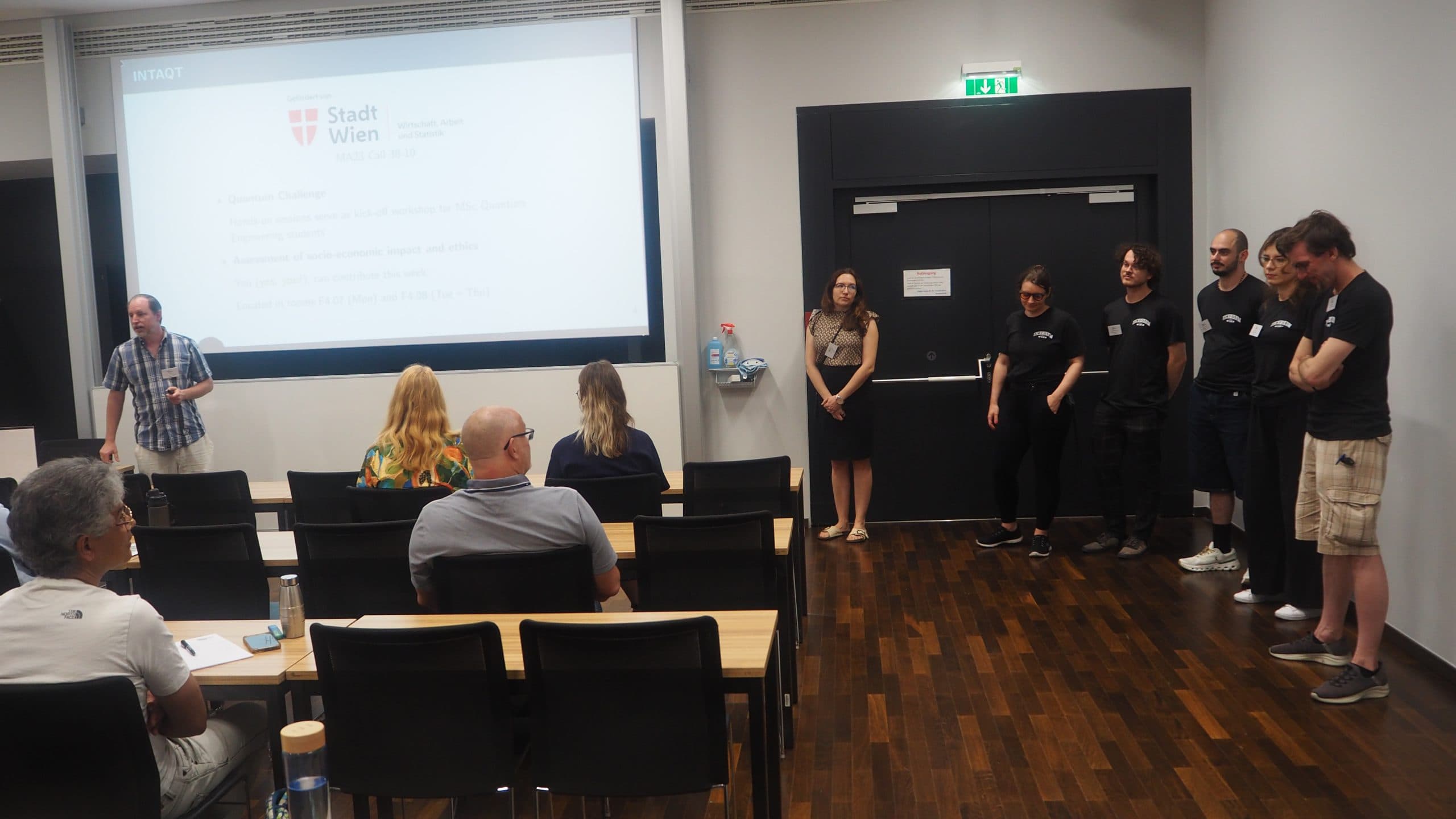 Eine Gruppe von Personen steht vor einem Klassenzimmer, einige andere sitzen vor einem Bildschirm, auf dem eine Präsentation mit dem Titel Stadt Wien gezeigt wird. Der Raum hat einen Holzboden und schwarze Türen an der Rückseite.