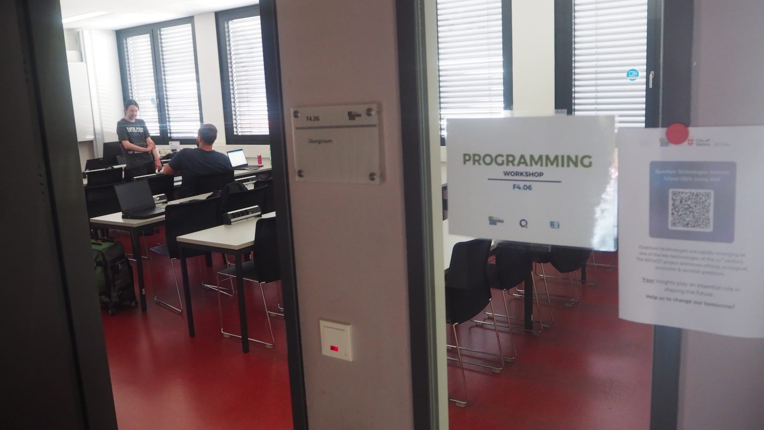 Ein Klassenzimmer mit mehreren Personen, die an Tischen sitzen, und einer Person, die vorne steht und spricht. Auf einem Schild an der Tür steht PROGRAMMIERWERKSTATT. Der Raum hat große Fenster und einen roten Bodenbelag.