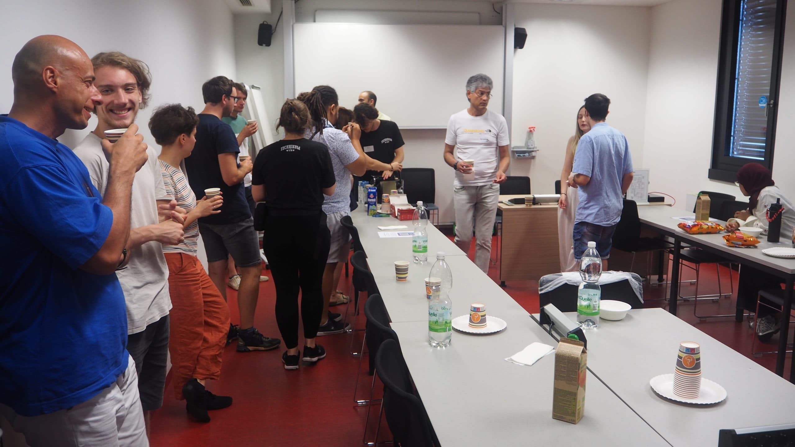 Eine Gruppe von Menschen sitzt in einem hellen Raum an Tischen mit Getränken und Snacks und unterhält sich, was auf ein zwangloses Treffen oder eine Büroveranstaltung hindeutet. Einige stehen, während andere sich von den Tischen aus bedienen.
