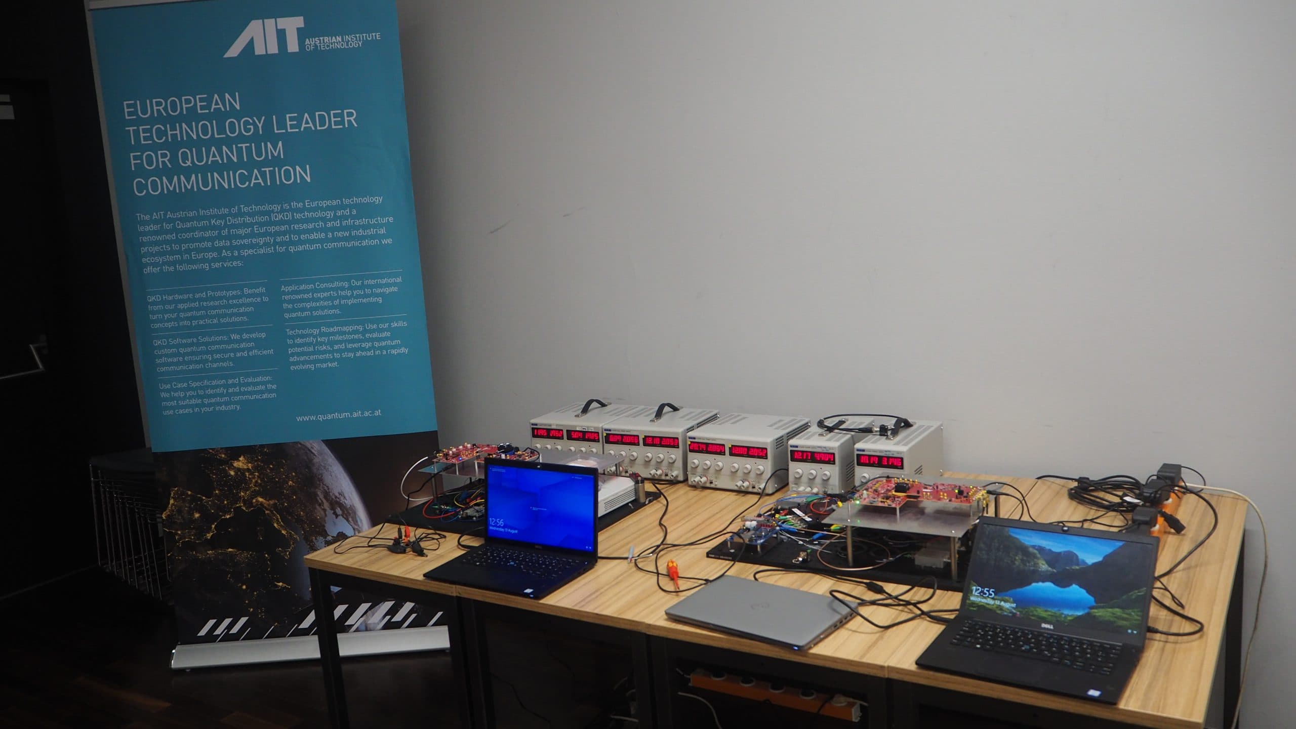 Ein Tisch mit Laptops, elektronischen Geräten und Kabeln, die für eine Demonstration der Quantenkommunikation aufgestellt sind, neben einem blauen Banner des AIT Austrian Institute of Technology, das auf die führende Rolle Europas in der Quantenkommunikation hinweist.