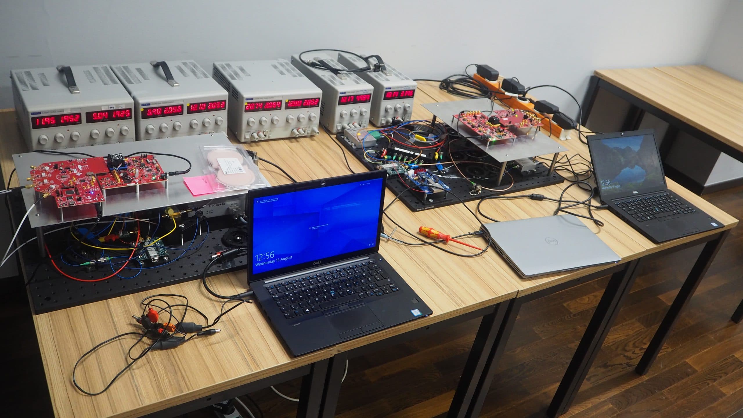Zwei Laptops auf einem Holztisch sind mit Leiterplatten und elektronischen Bauteilen verbunden, im Hintergrund sind drei digitale Netzteile zu sehen. Drähte und Werkzeuge liegen verstreut um die Geräte herum.