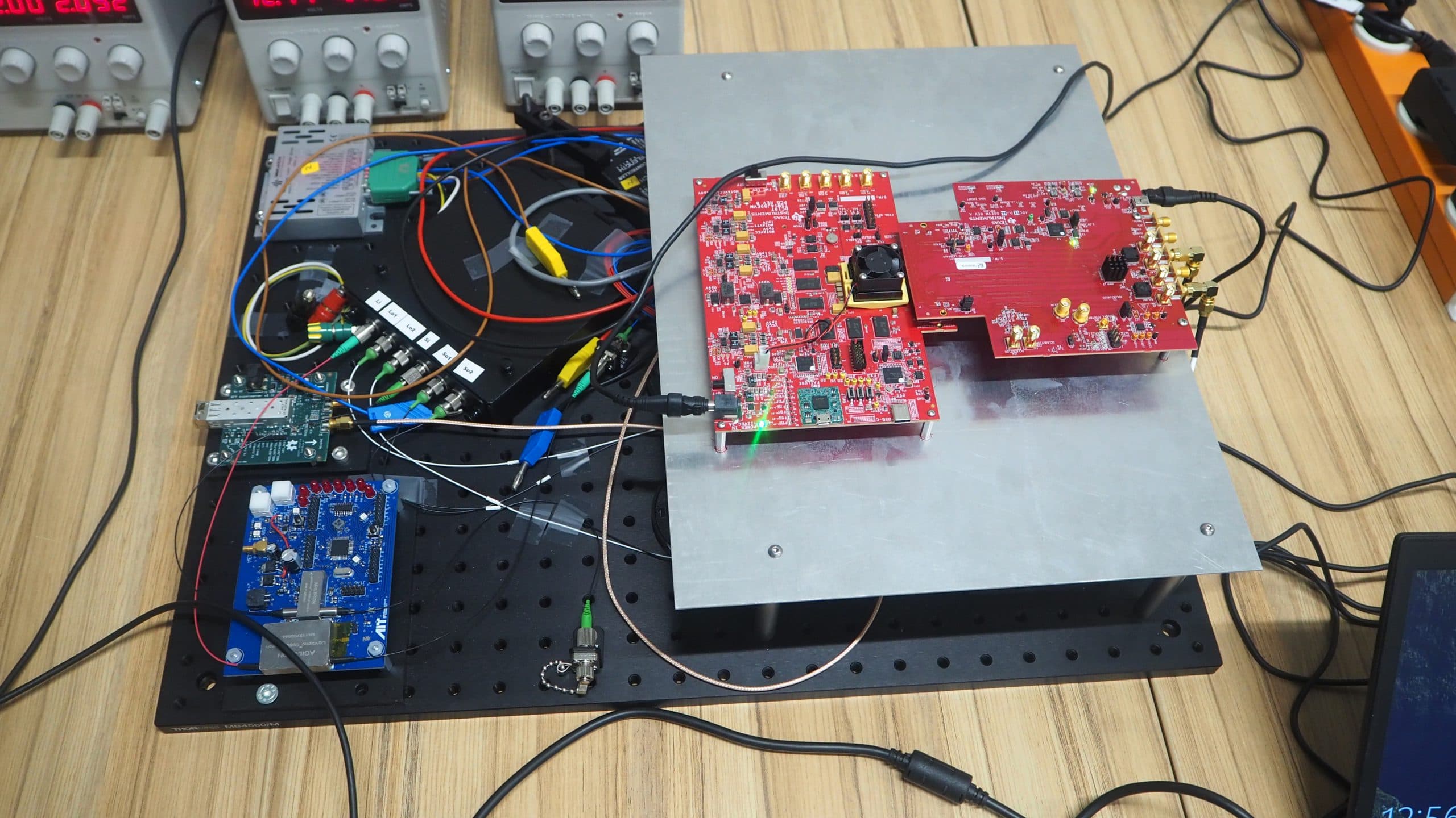 Ein Elektronik-Tischaufbau besteht aus mehreren Platinen, darunter eine große rote Platine mit angeschlossenen Kabeln, eine blaue Platine und verschiedene angeschlossene Komponenten und Drähte, die alle auf einer perforierten schwarzen Plattform stehen.
