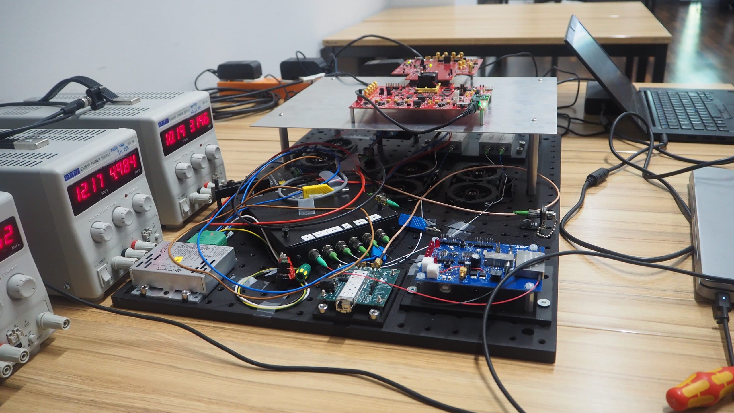 Ein Elektroniklabor auf einem Holztisch mit offenen Leiterplatten, verwickelten Drähten, Messgeräten, die Spannungen anzeigen, und einem angeschlossenen Laptop. Mehrere Netzteile und elektronische Bauteile sind zu sehen.