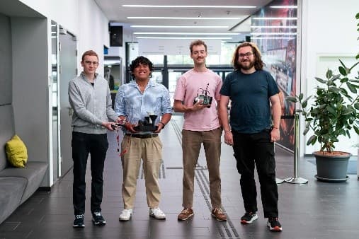 Vier Personen stehen in einem modernen Hausflur, zwei von ihnen halten kleine Robotergeräte. Sie sind lächelnd und leger gekleidet, im Hintergrund sind Pflanzen und Sitzgelegenheiten zu sehen.