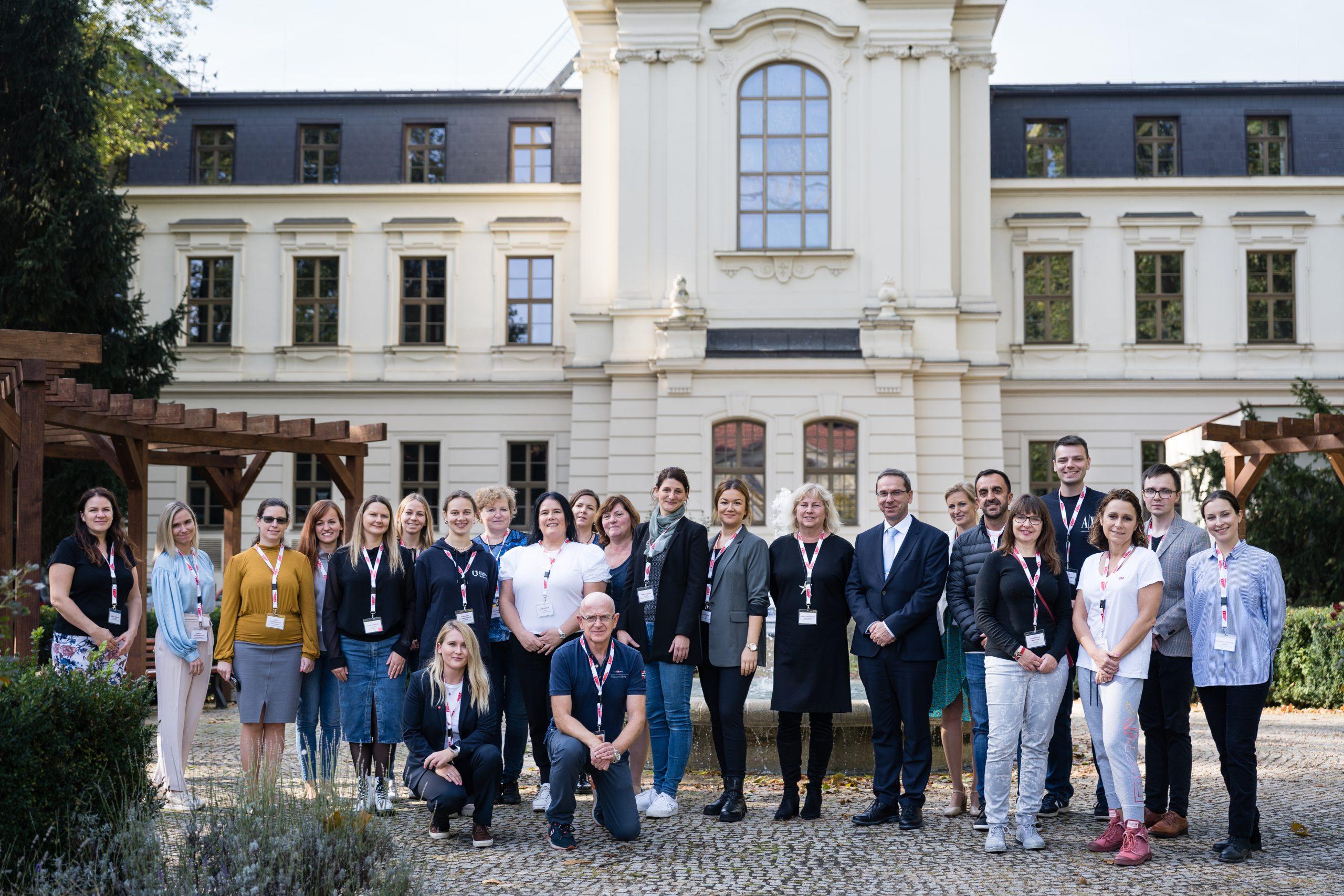 Eine Gruppe von etwa 25 Personen, Männer und Frauen, steht vor einem großen, eleganten Gebäude mit weißen Wänden und Bogenfenstern. Die meisten von ihnen tragen legere Geschäftskleidung und Namensschilder und posieren im Freien auf einem Kopfsteinpflaster.