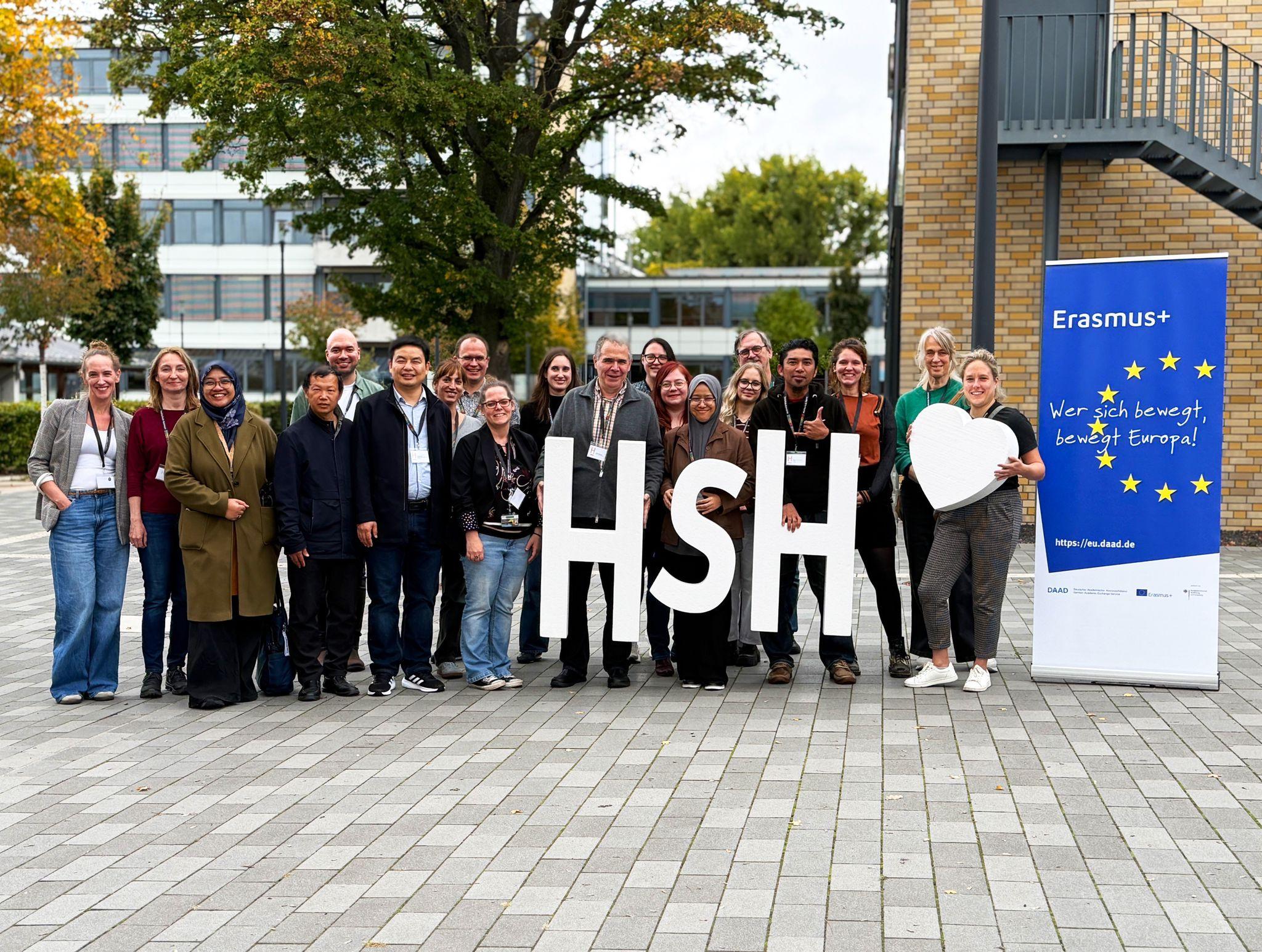 Eine Gruppe von Menschen steht im Freien, lächelt und hält große Buchstaben mit der Aufschrift HSH und ein weißes Herz in der Hand. Rechts ist ein blaues Erasmus+-Banner zu sehen, und im Hintergrund sind Bäume und Gebäude zu sehen.