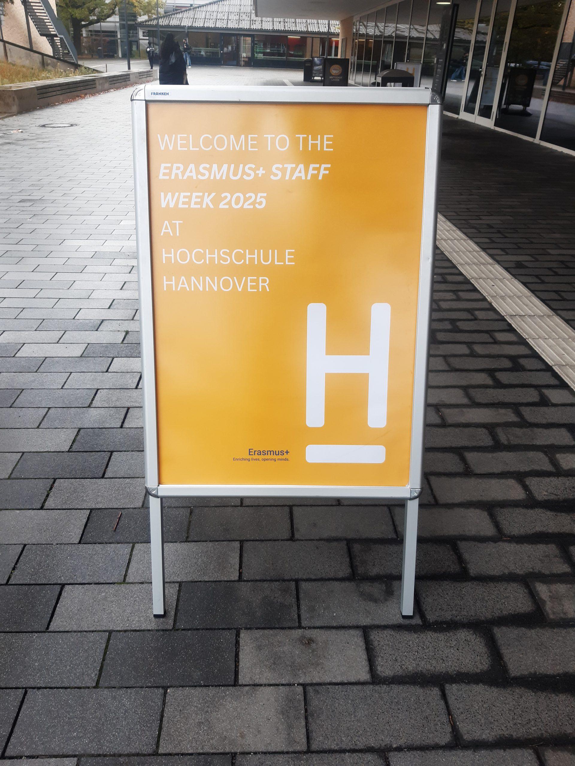 Auf einem gelben Schild auf einem gepflasterten Gehweg steht: Willkommen zur Erasmus+ Staff Week 2025 an der Hochschule Hannover in weißer und helloranger Schrift. Das Logo der Hochschule Hannover befindet sich am unteren Rand.